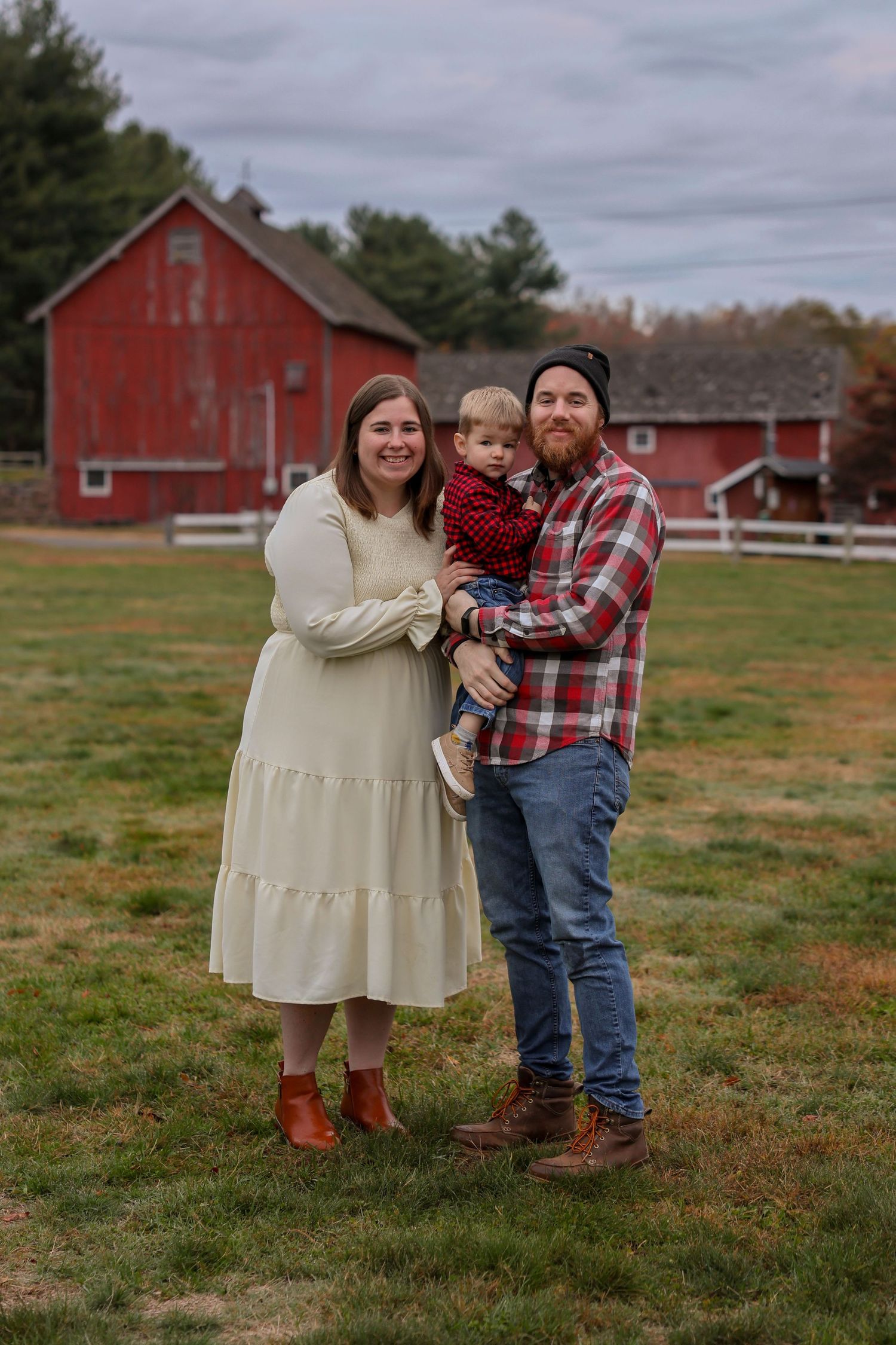 Young family with toddler poses in front of red barn during fall family photo session.