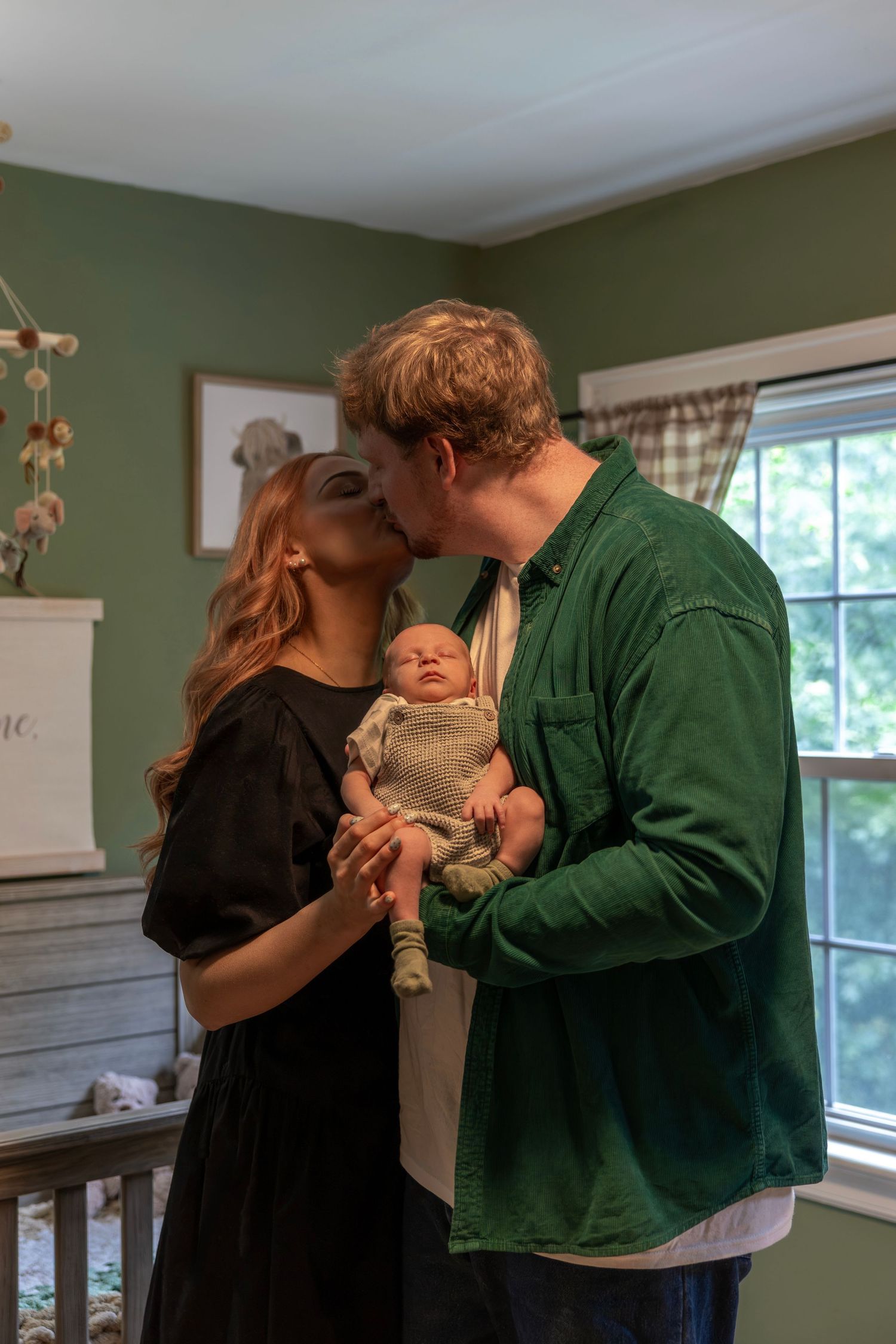 A couple shares a kiss while holding their newborn baby in a cozy green nursery room.