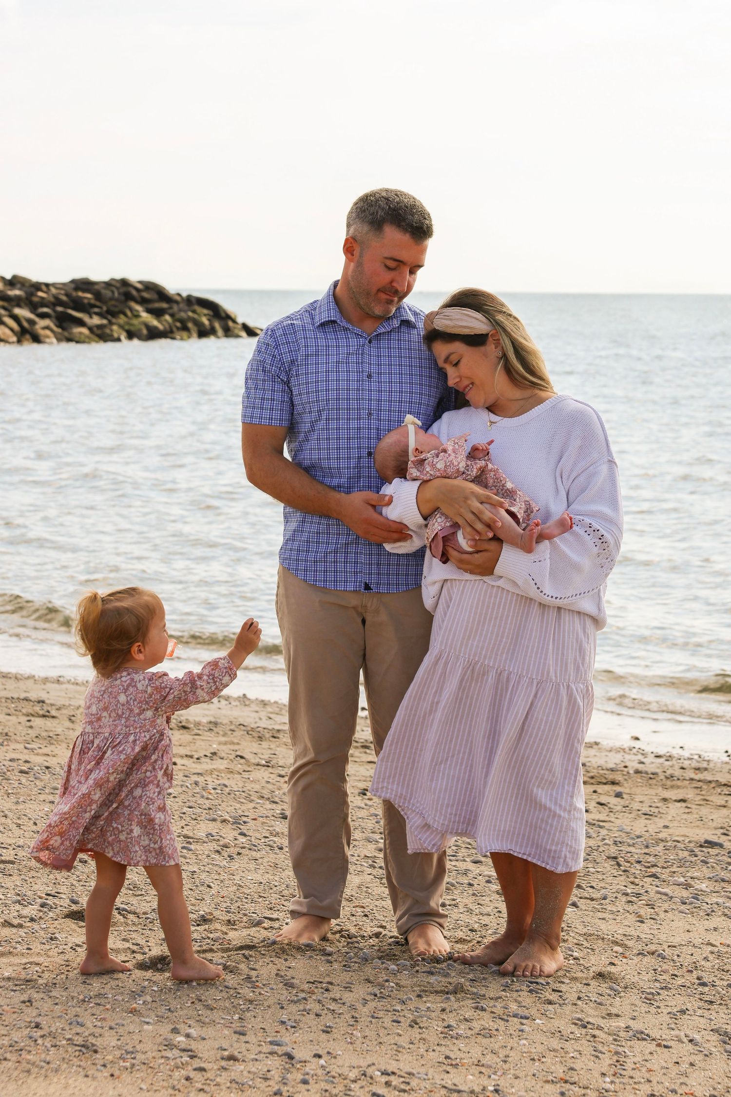 Family of four with newborn and toddler daughter share tender moment together on rocky beach.