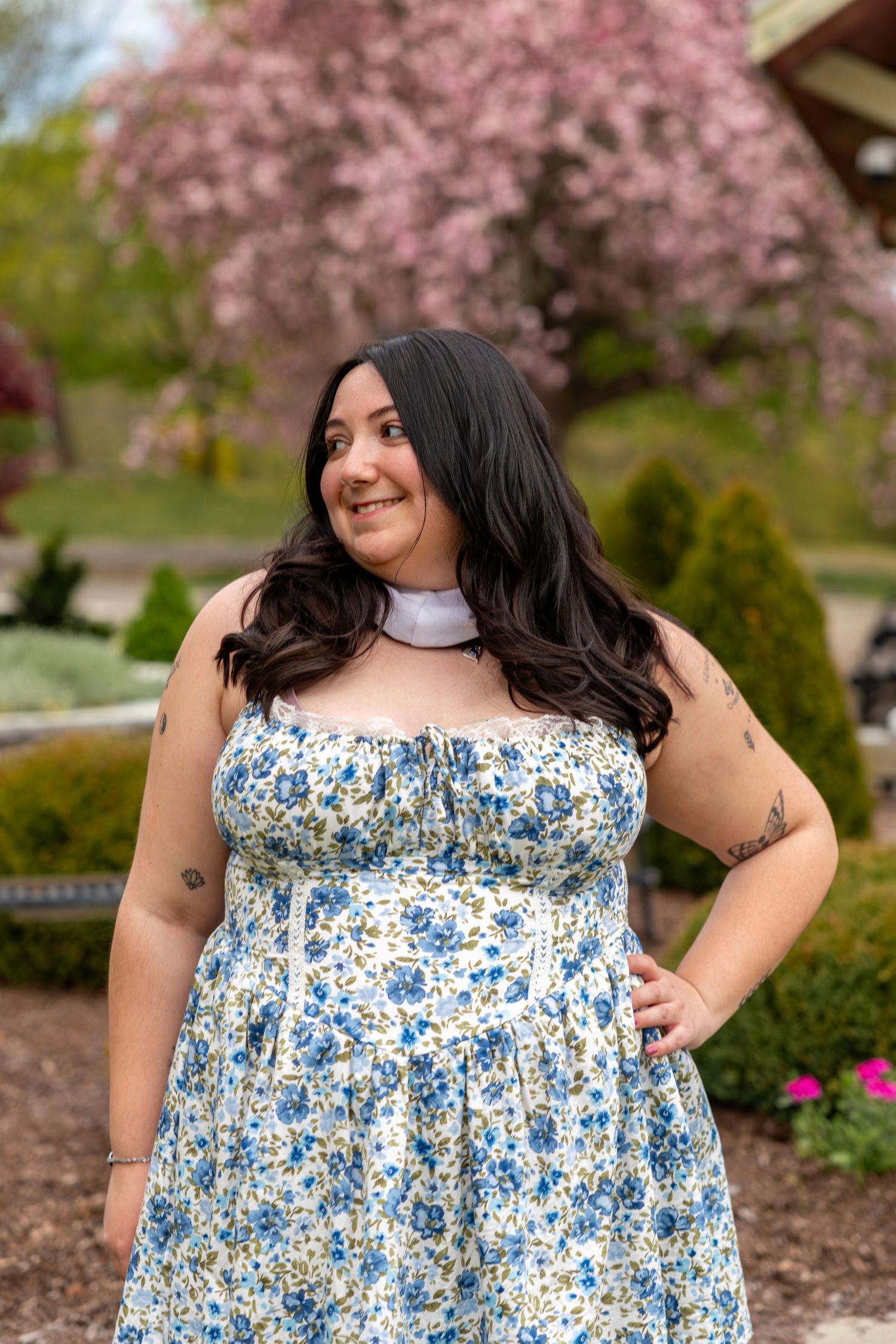 Young woman with tattoos in blue floral strapless dress smiles in blooming spring garden with pink cherry trees.