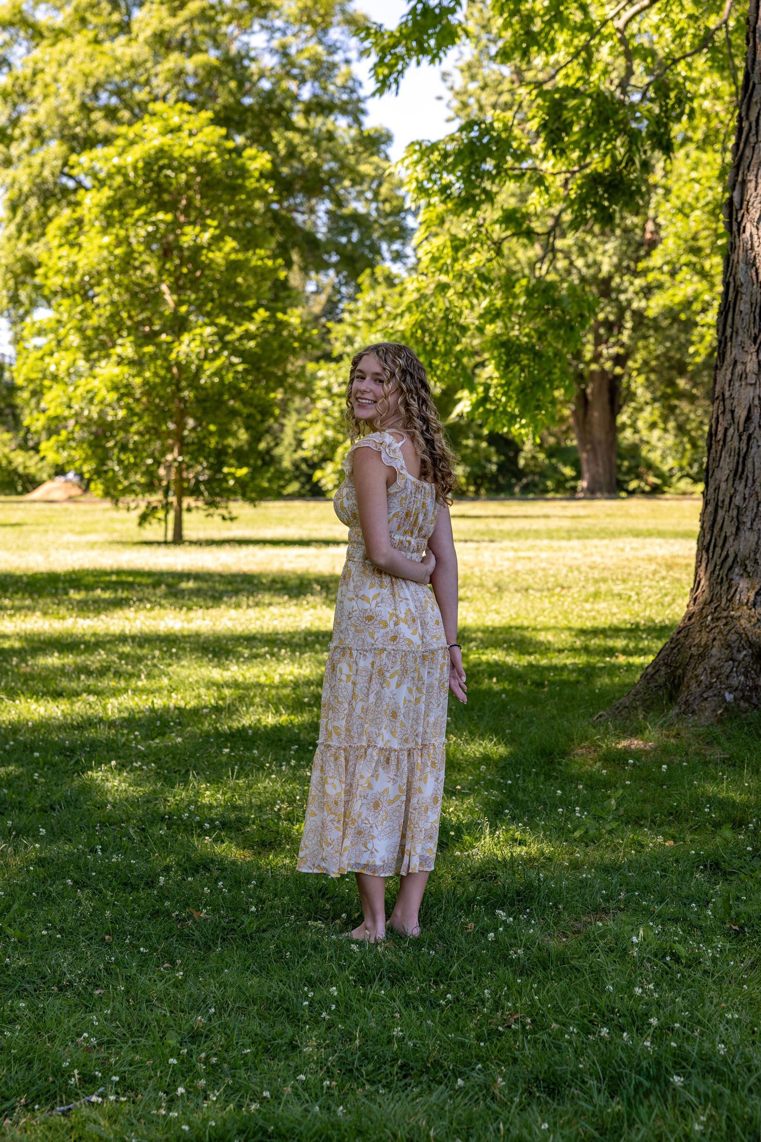 Young woman in floral midi dress standing barefoot on lush green grass beside large tree.