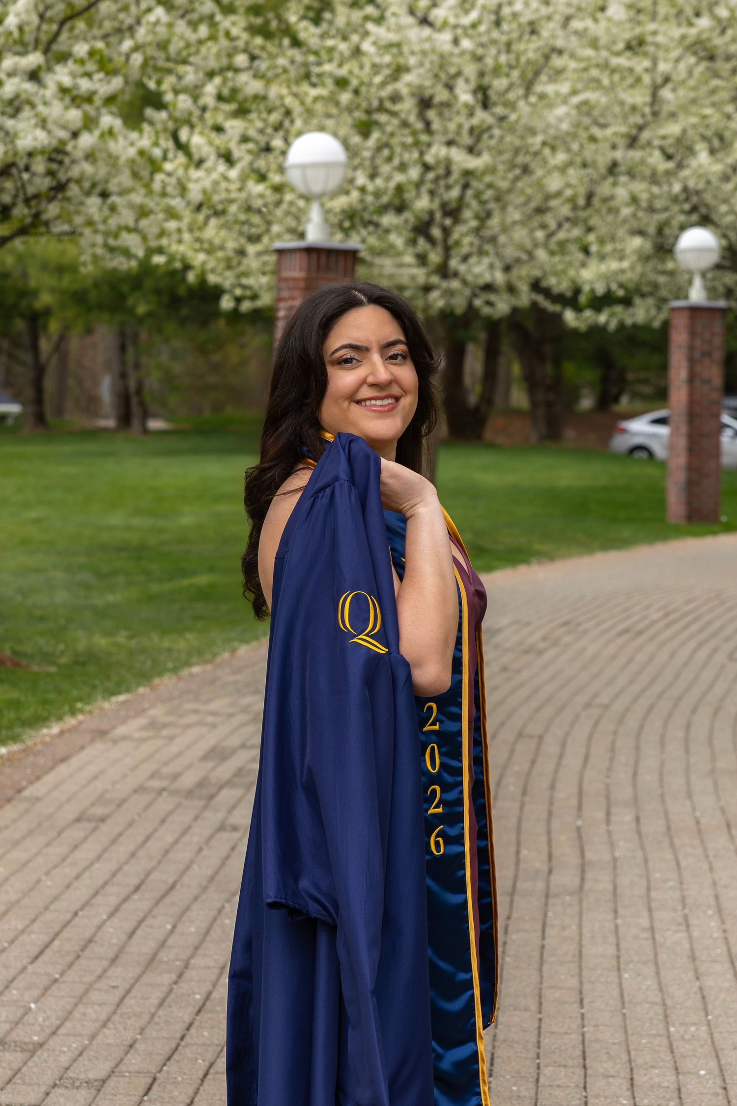 Female graduate holding navy blue Quinnipiac University gown near blooming white flowering trees.