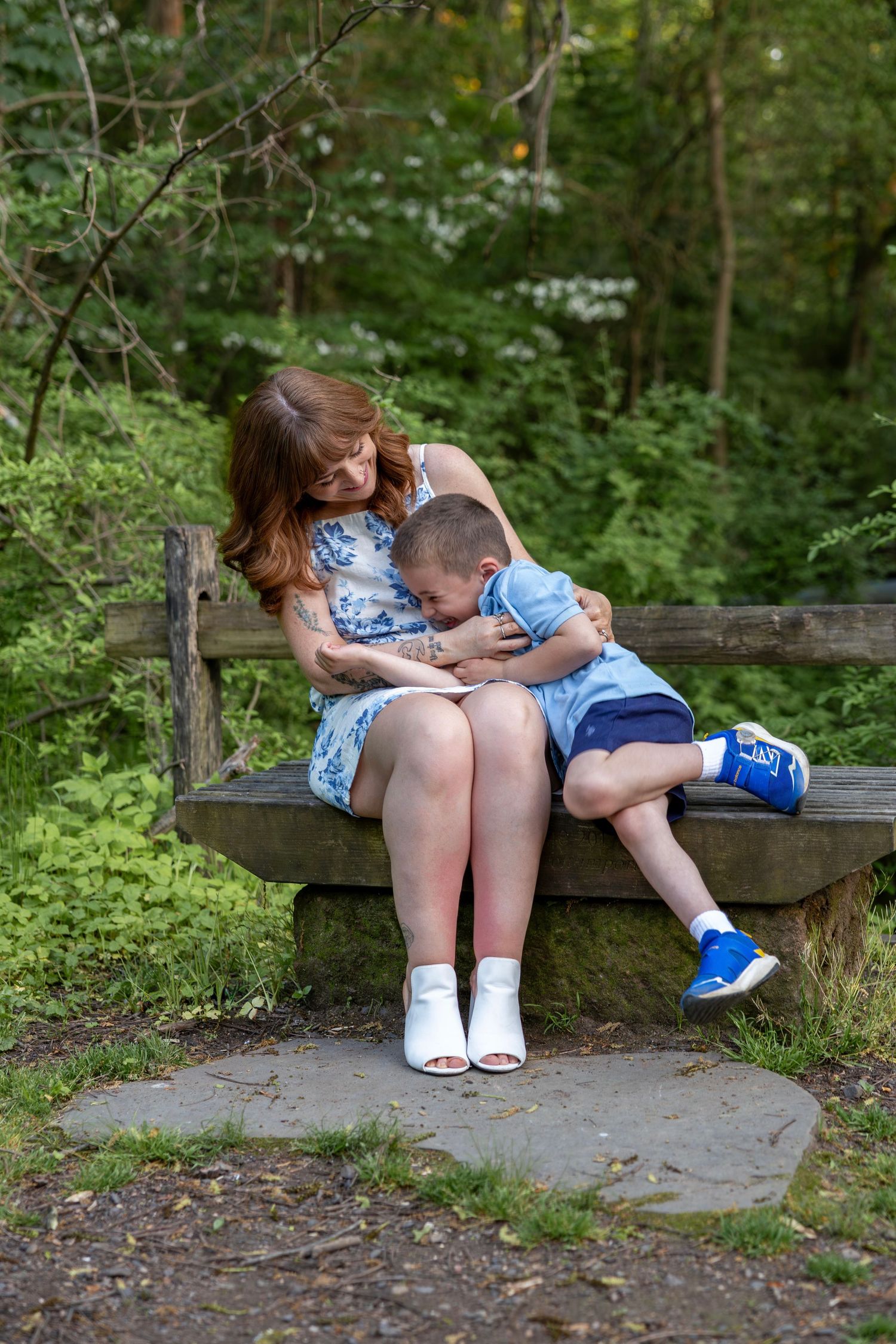 Tender moment as mother and young son sit closely together on a wooden bench surrounded by green foliage.
