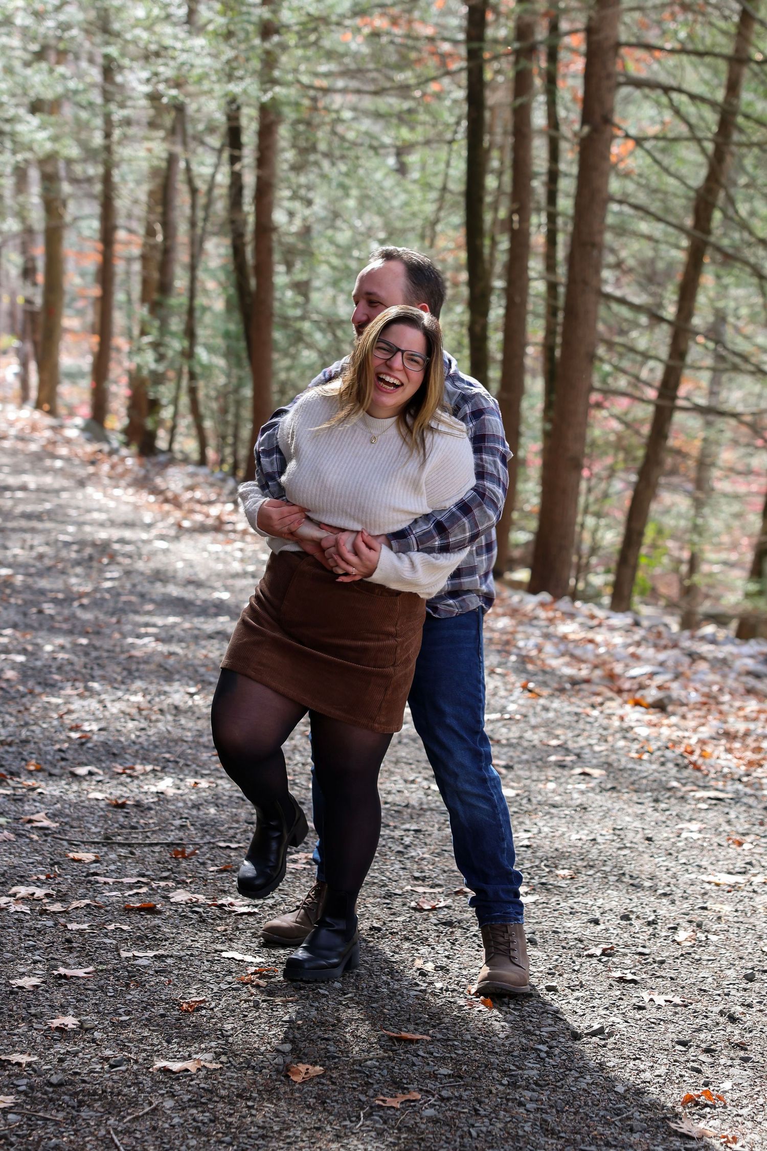 A laughing couple playfully embraces on a wooded gravel path surrounded by tall trees in autumn.