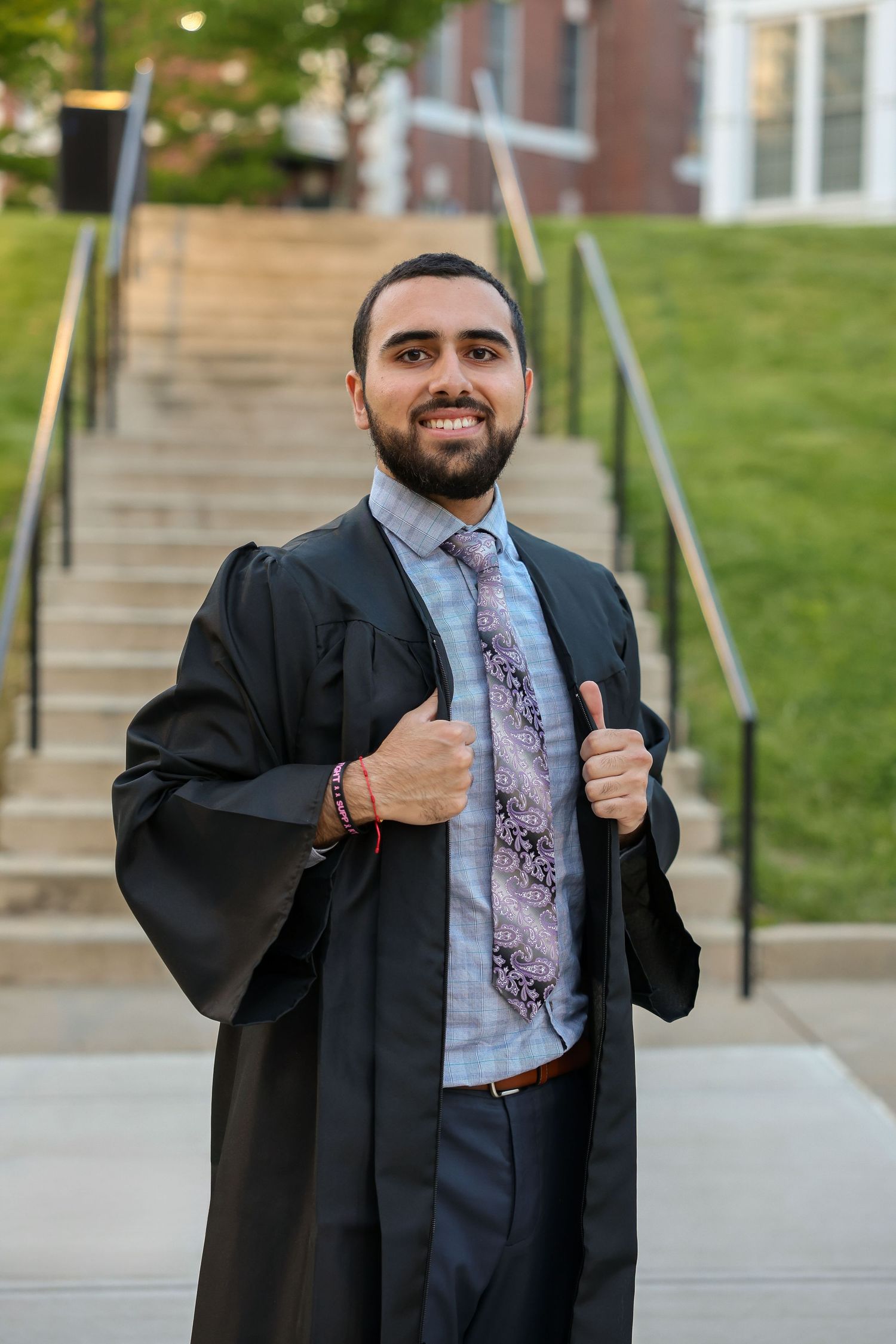 Male graduate in black gown gives thumbs up wearing floral tie in front of brick campus steps.