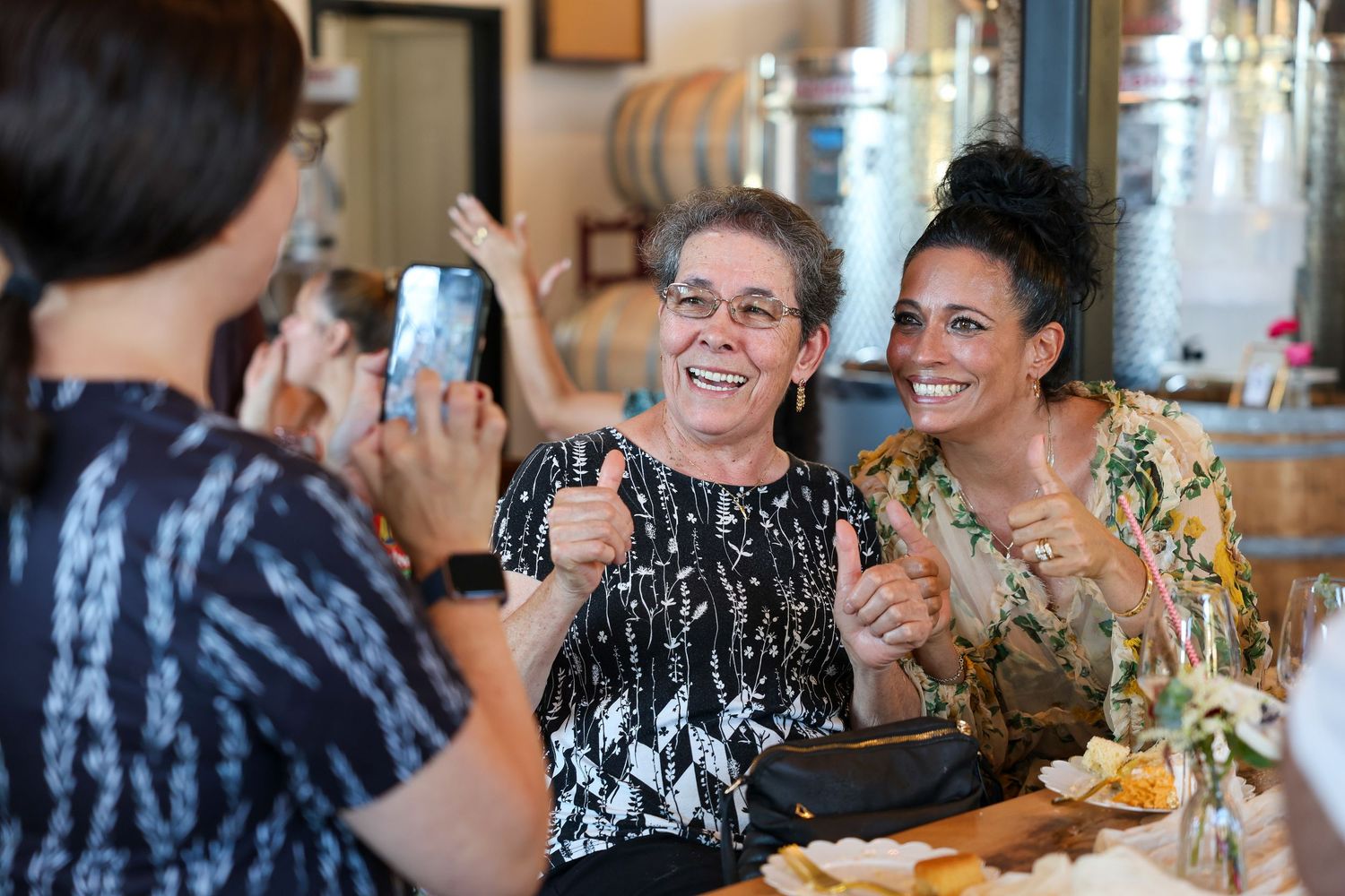 Two women giving thumbs up while being photographed at a lively indoor winery celebration.