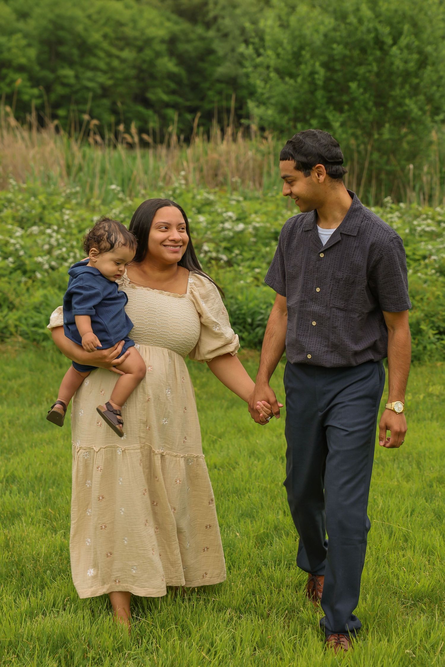 Pregnant woman carries toddler while holding hands with partner walking in green field.