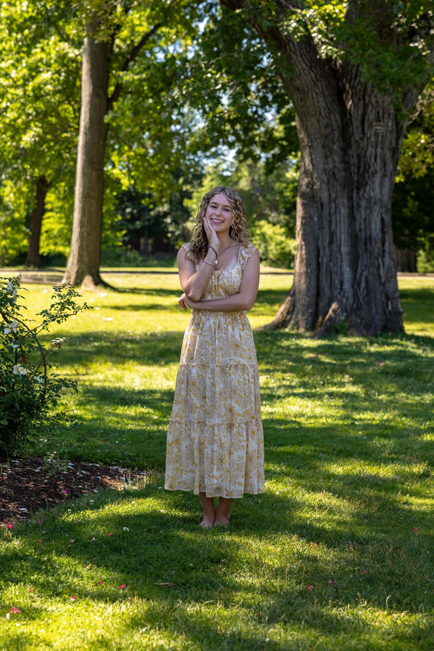 Young woman in yellow floral maxi dress standing barefoot on green grass in a sunny park with large trees.