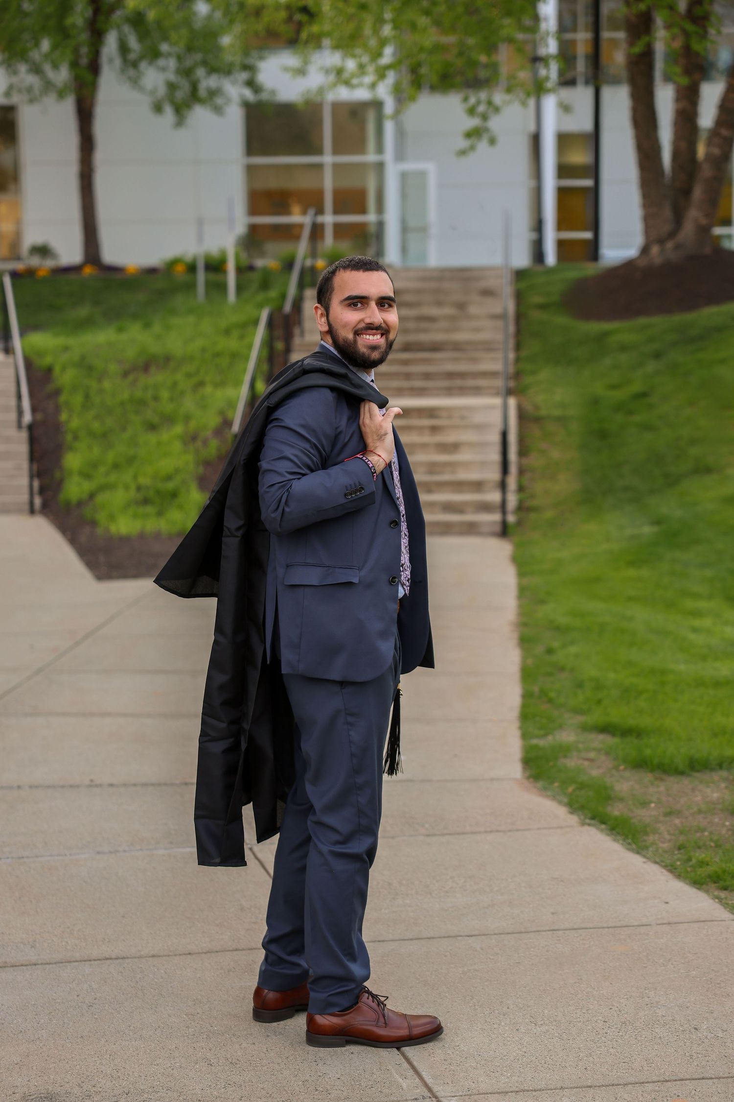 Young man in navy blue suit carrying jacket over shoulder on college campus walkway.