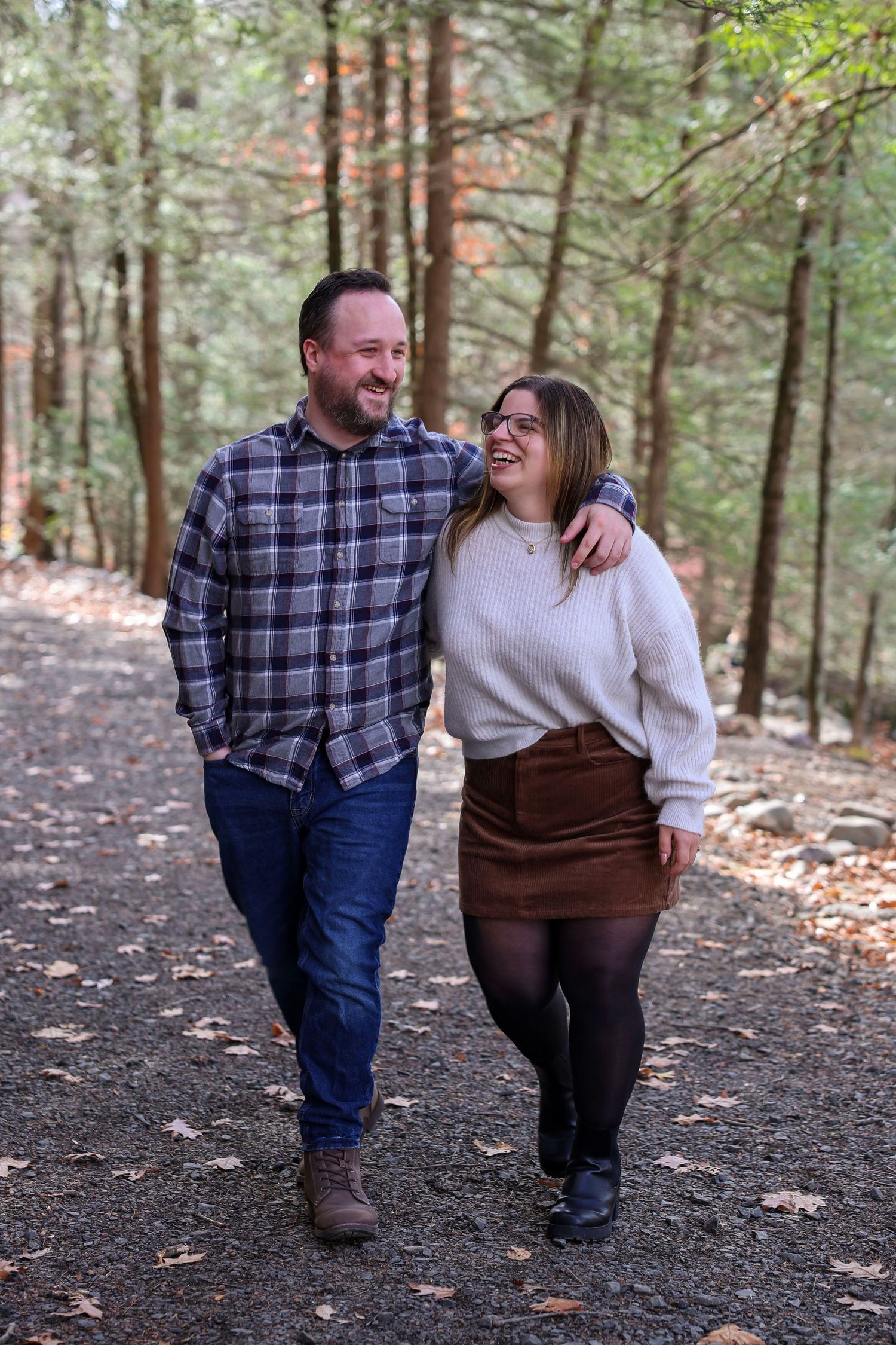 A happy couple walks arm-in-arm along a forest trail, laughing together among tall autumn trees.