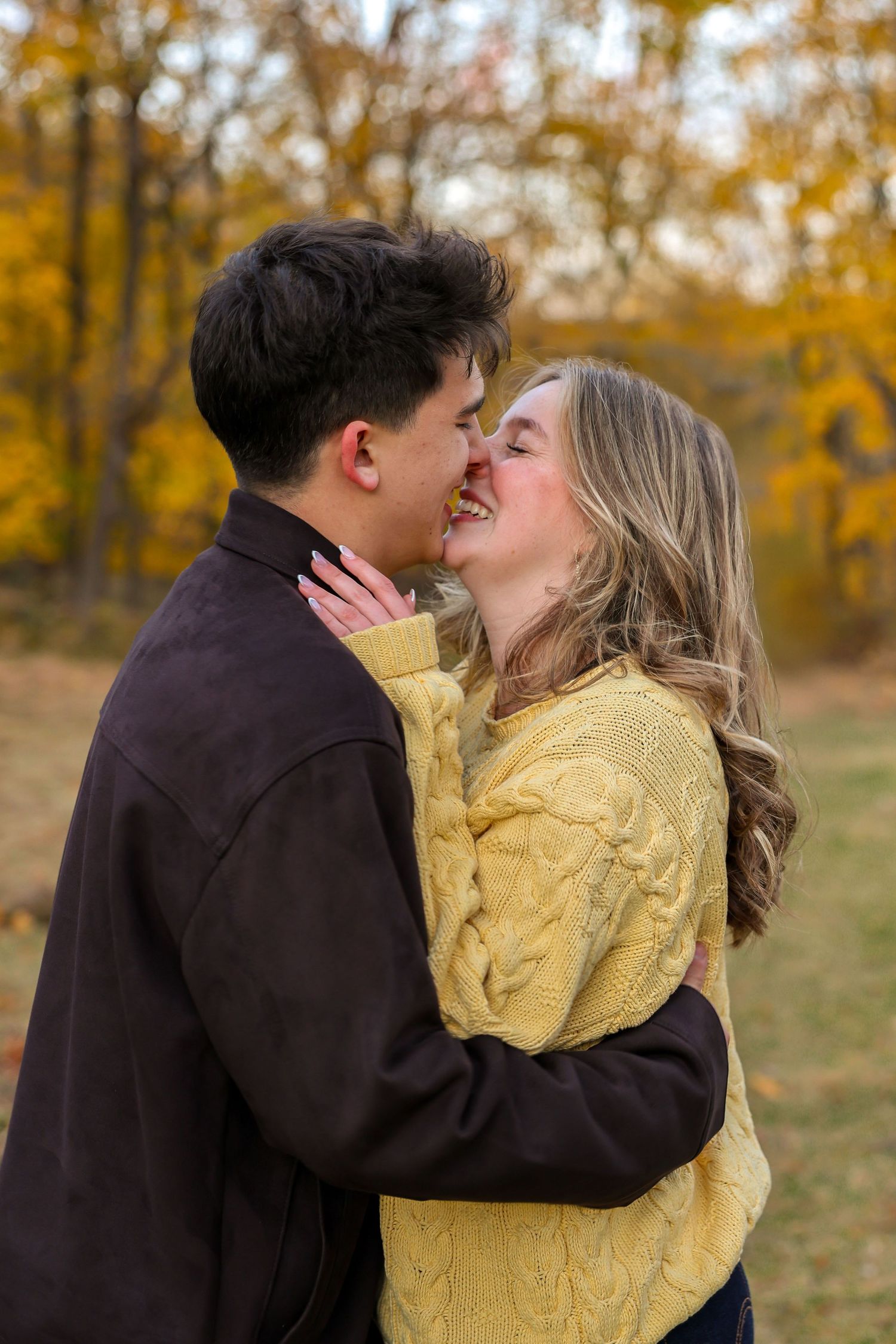 A joyful young couple sharing a kiss and laughing together amid vibrant autumn foliage outdoors.