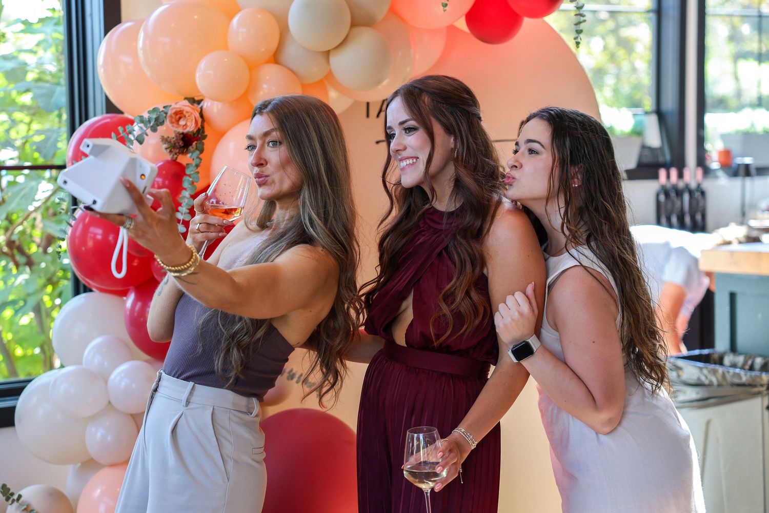 Three women posing for selfie at elegant party with balloon arch backdrop.