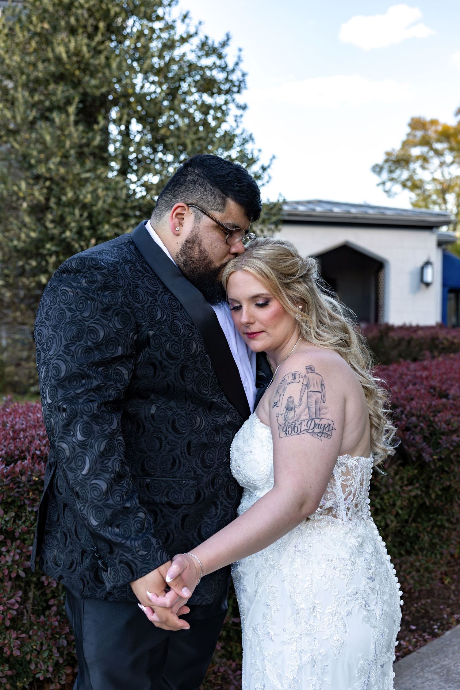 Groom in black floral tuxedo kissing bride on head while holding hands outdoors at wedding venue.