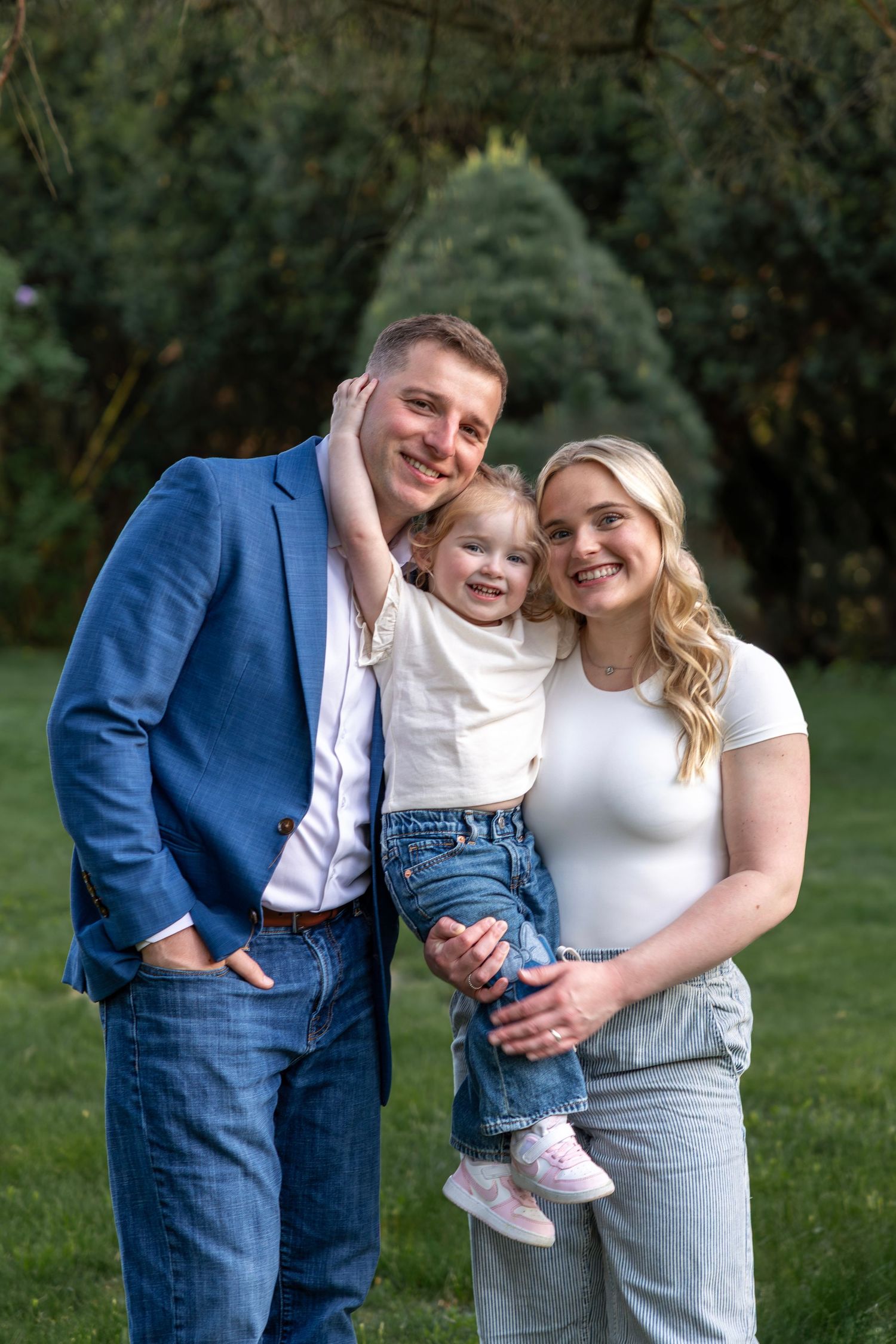 Happy family of three posing outdoors, parents holding toddler in garden setting with green trees background.