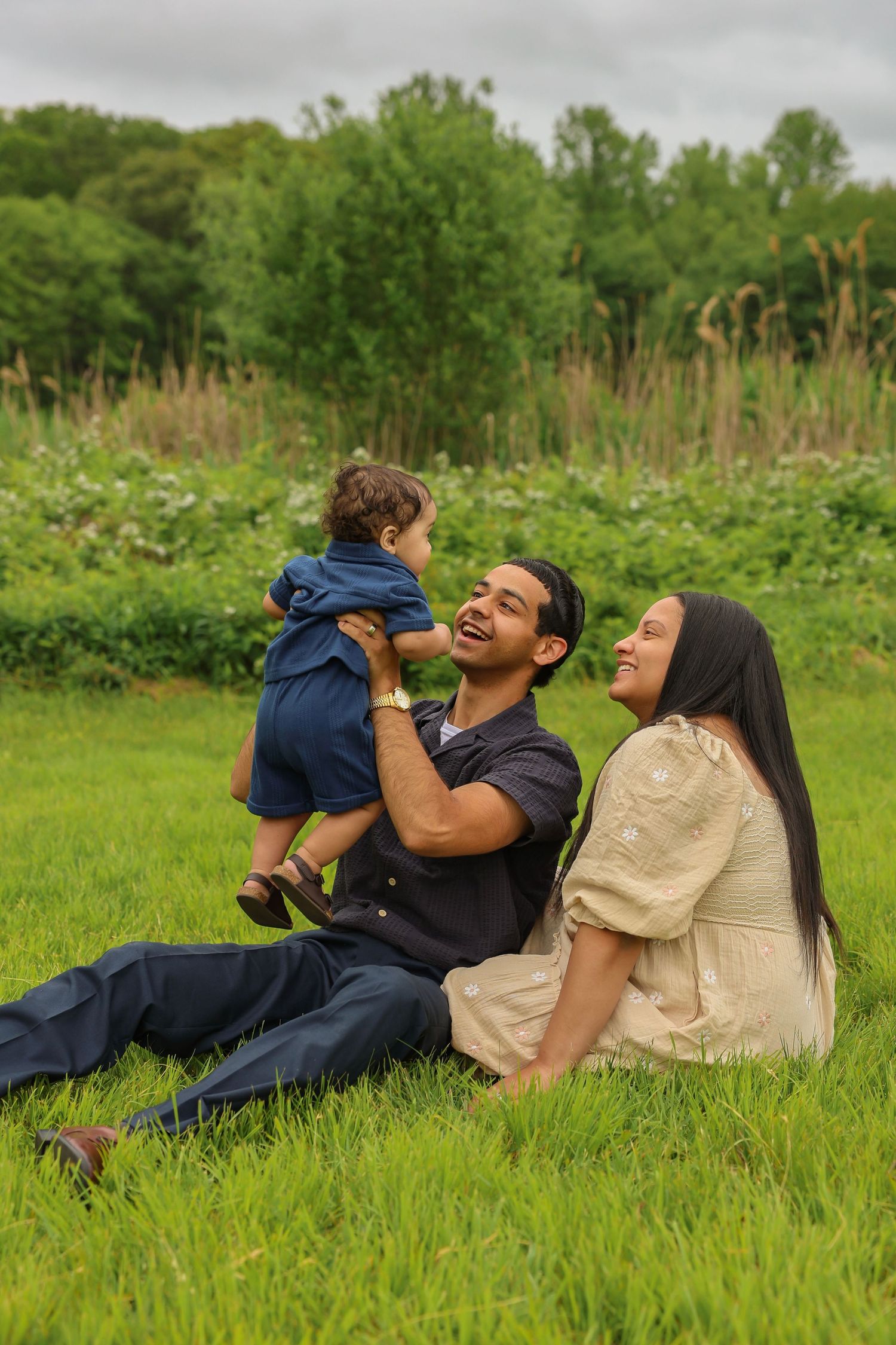 Laughing father lifts baby in air while mother watches joyfully on green grass.