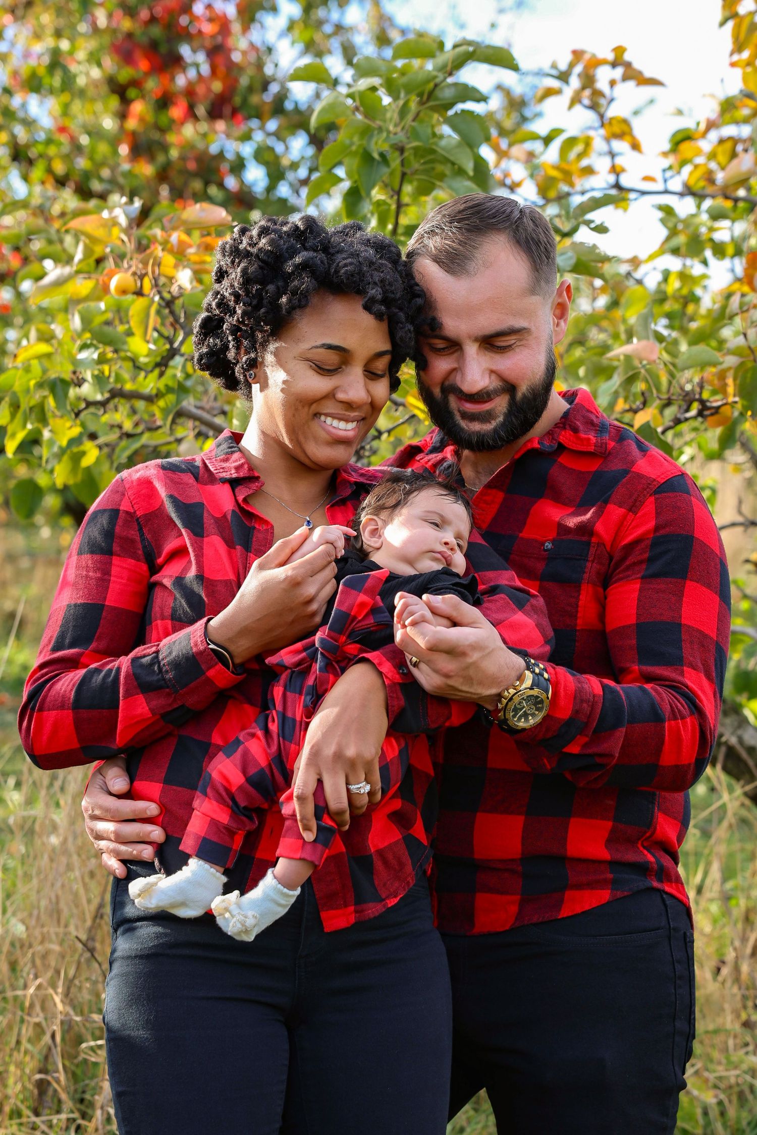 Interracial family in matching red plaid shirts smiling outdoors in autumn orchard while holding their newborn baby.