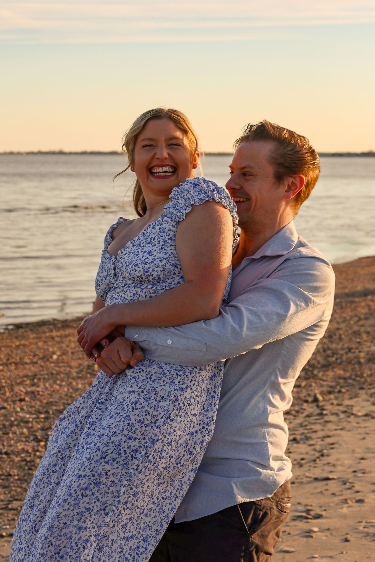 Joyful couple laughing and embracing on a sandy beach during a warm sunset.