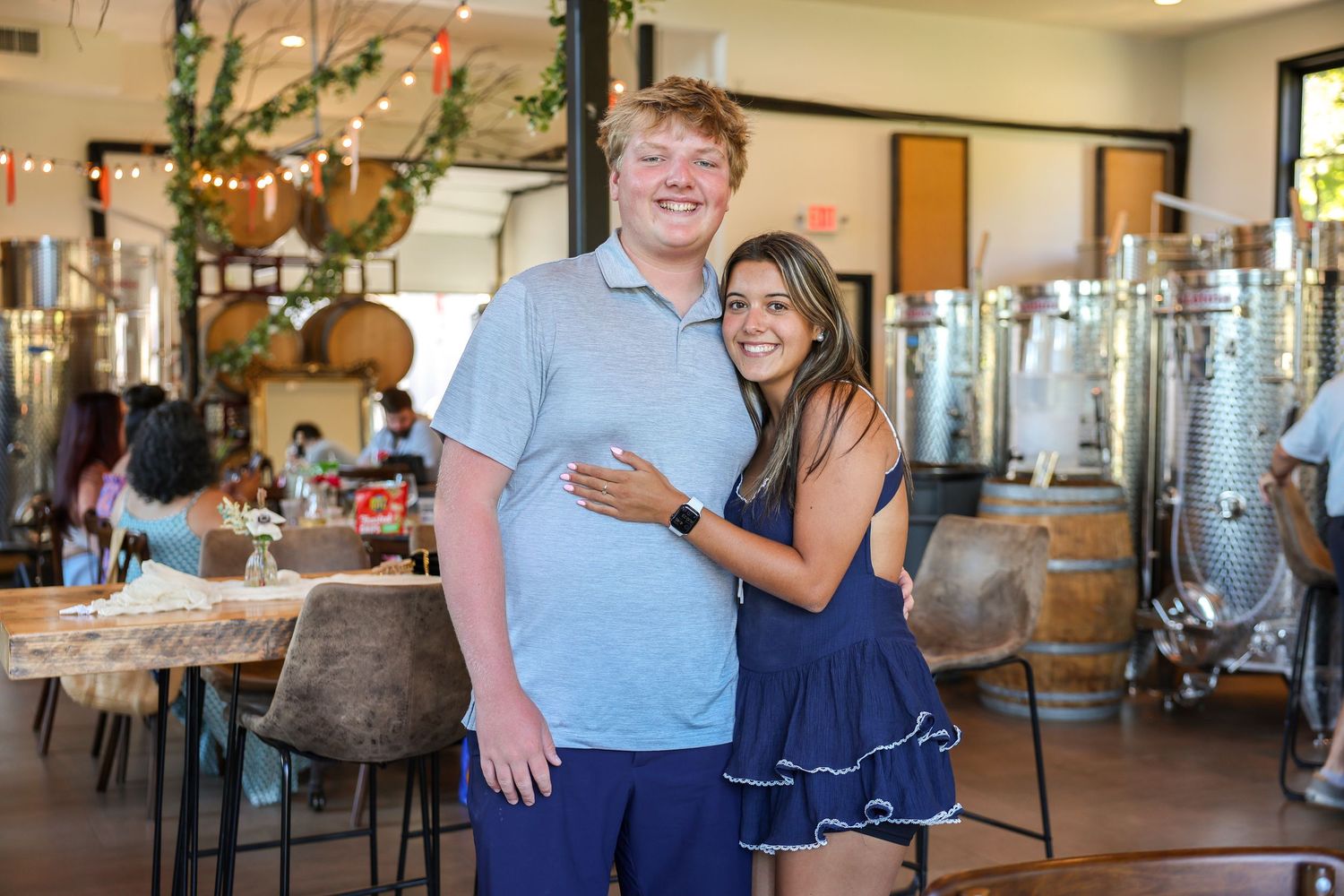 Young couple smiling and posing together inside a modern winery with steel tanks in background.