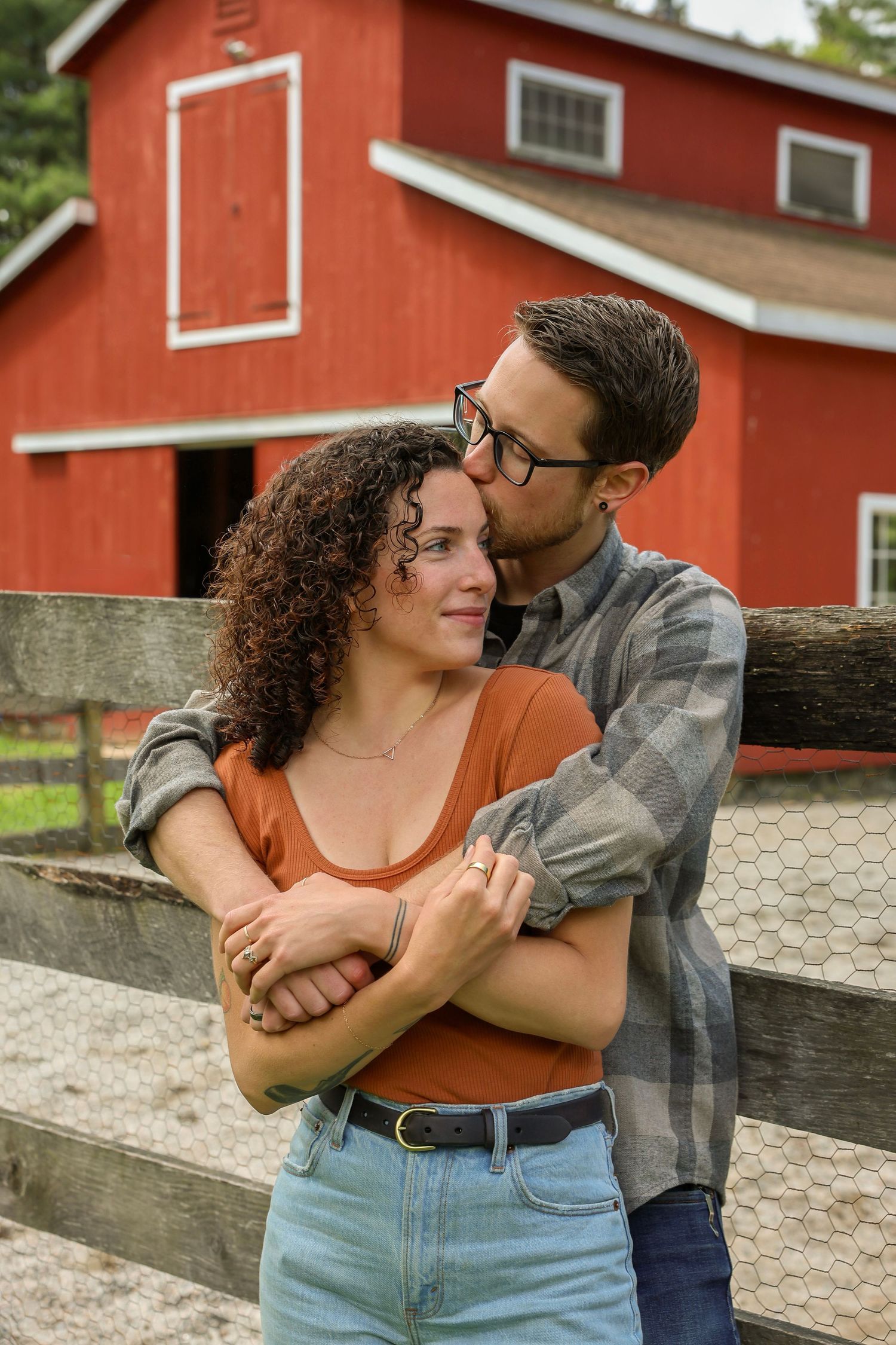 A romantic couple embracing near a wooden fence with a red barn backdrop on a farm.