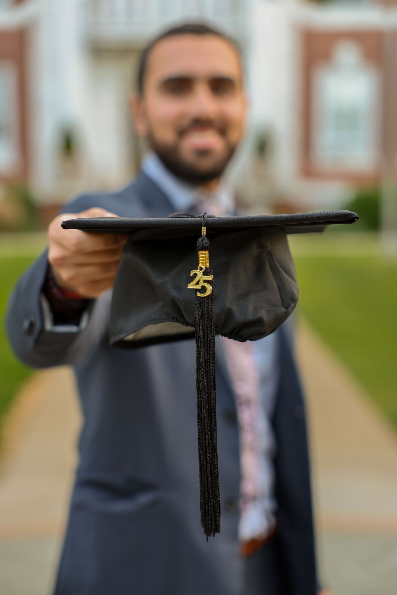 Male graduate holds black mortarboard cap with gold 25 tassel toward camera on campus pathway.