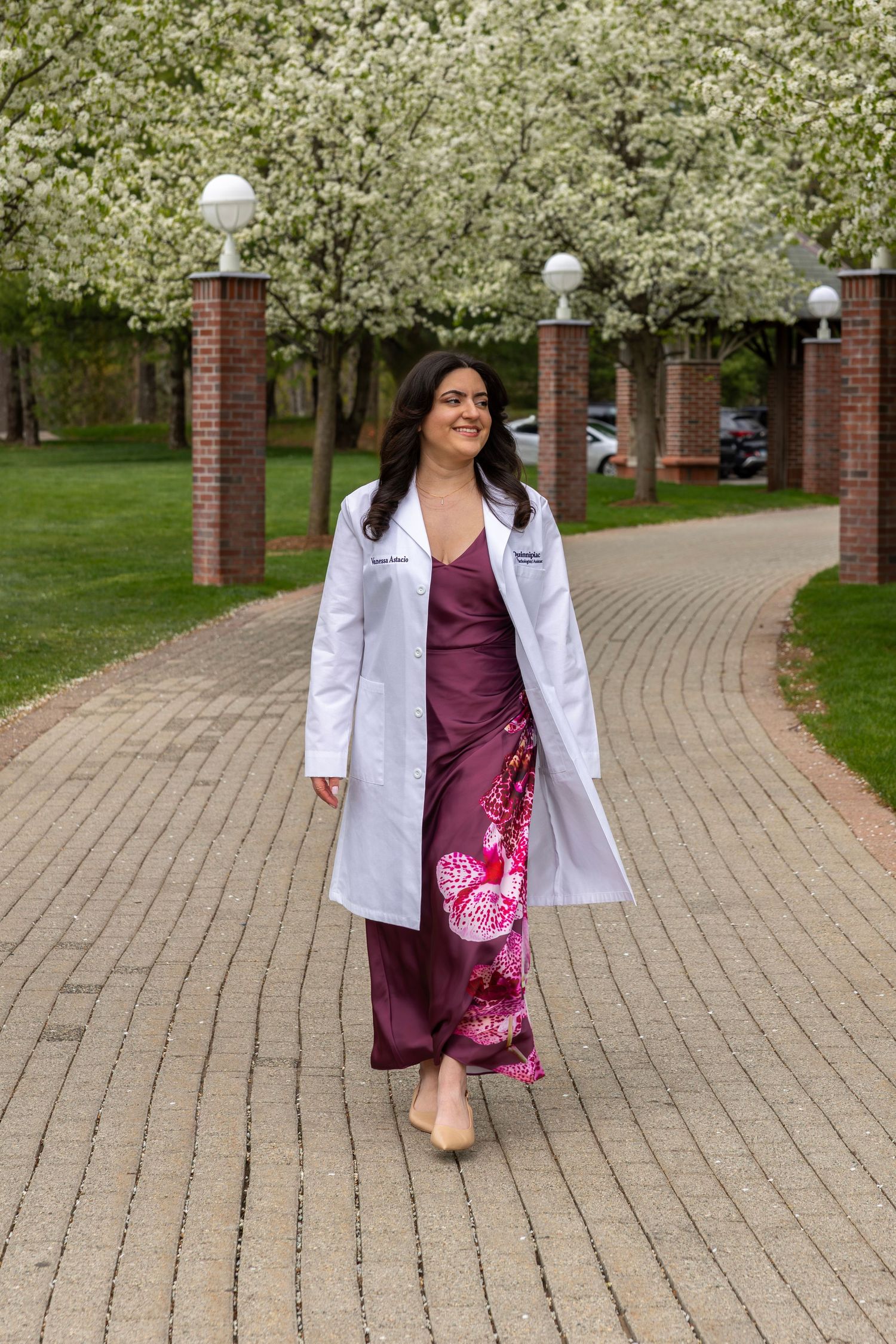 Female medical graduate in white coat and burgundy dress walking among blooming white trees.