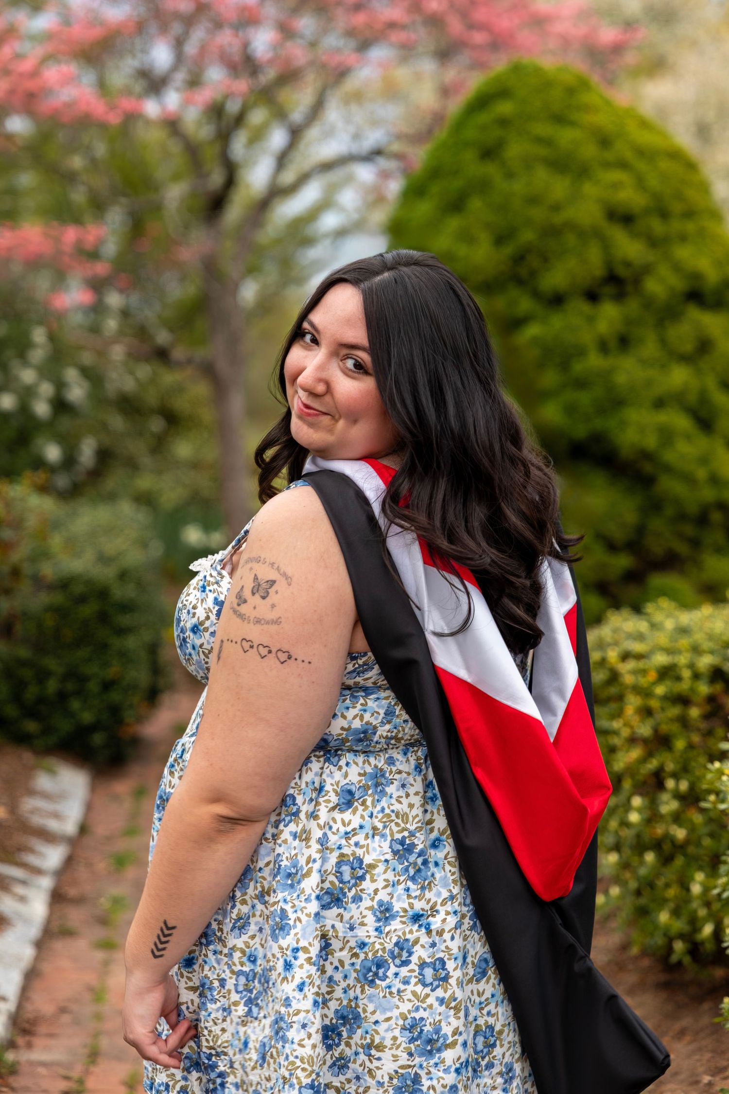 Smiling woman in floral dress wearing graduation regalia poses in a blooming garden during graduation celebration.