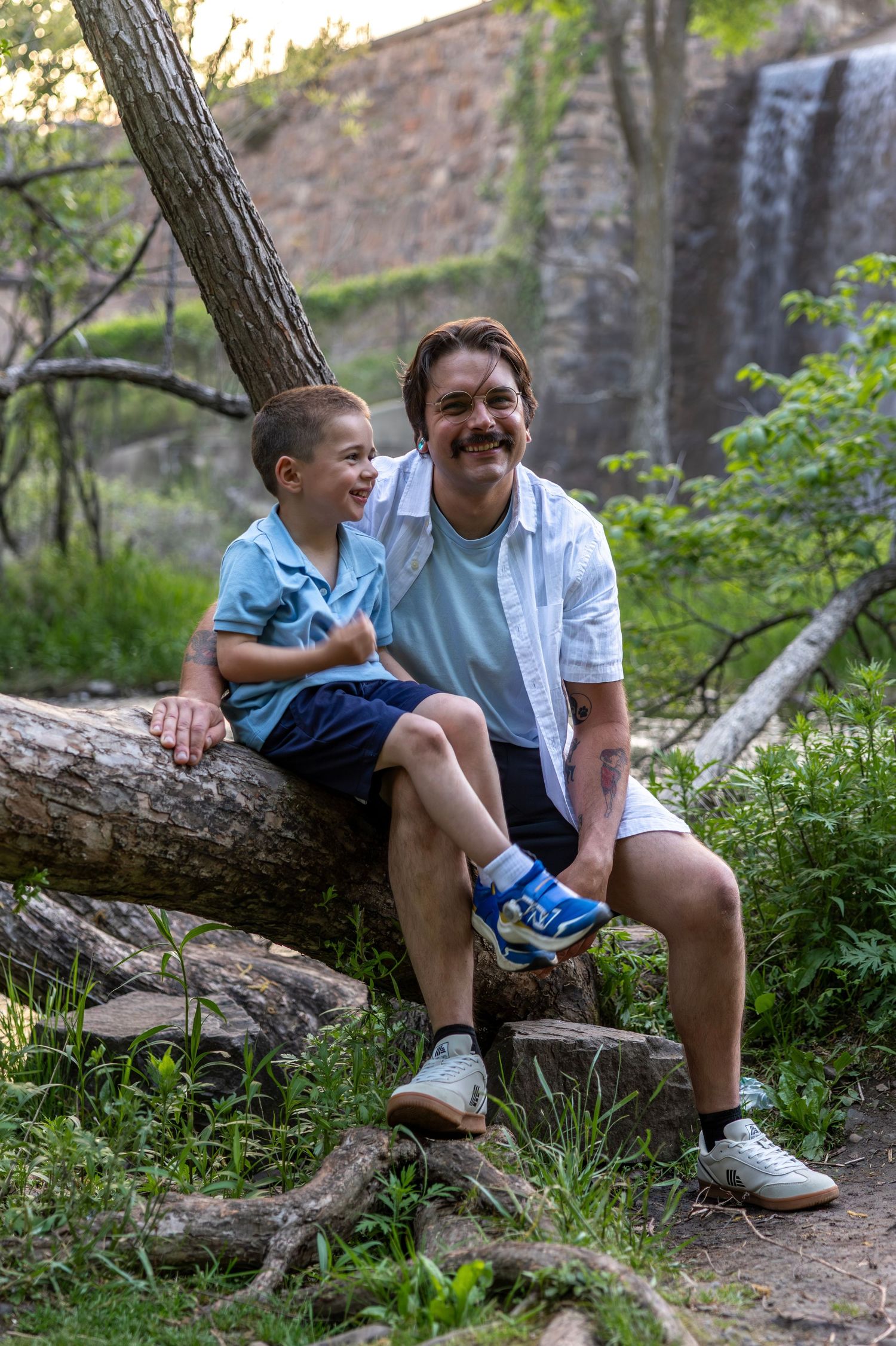 Father and young son sit together on a fallen log laughing near a scenic waterfall in a lush forest.