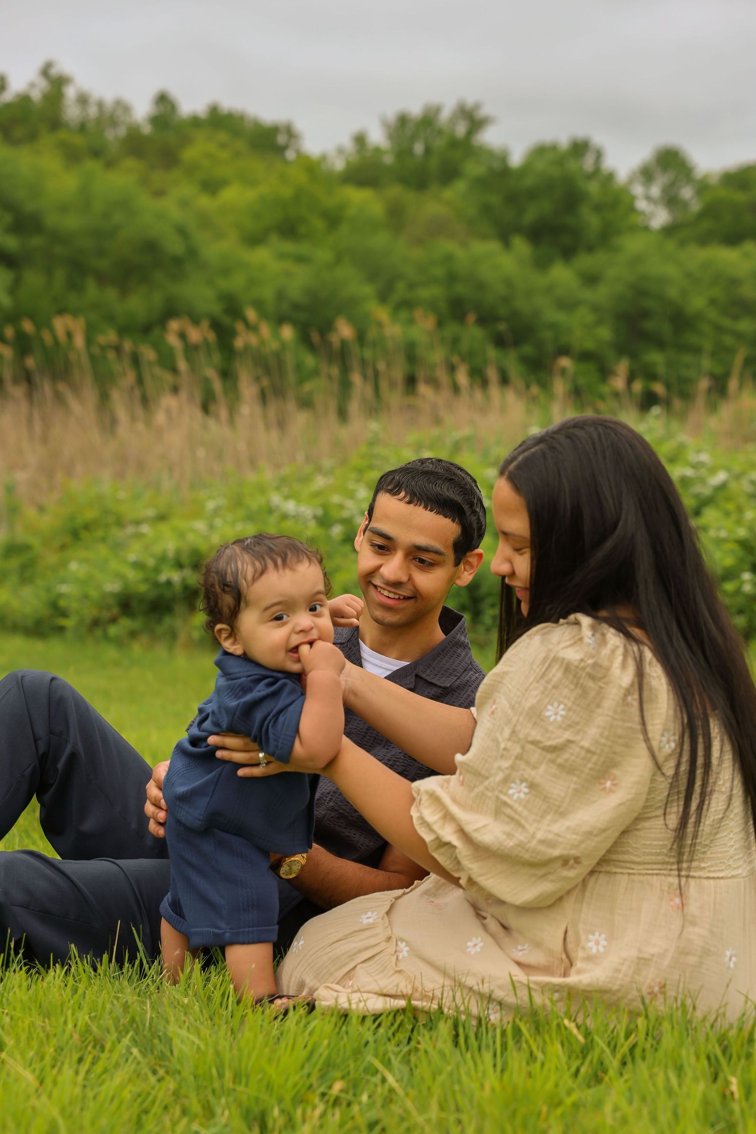 Happy family sits in grass as parents smile at baby in blue outfit.