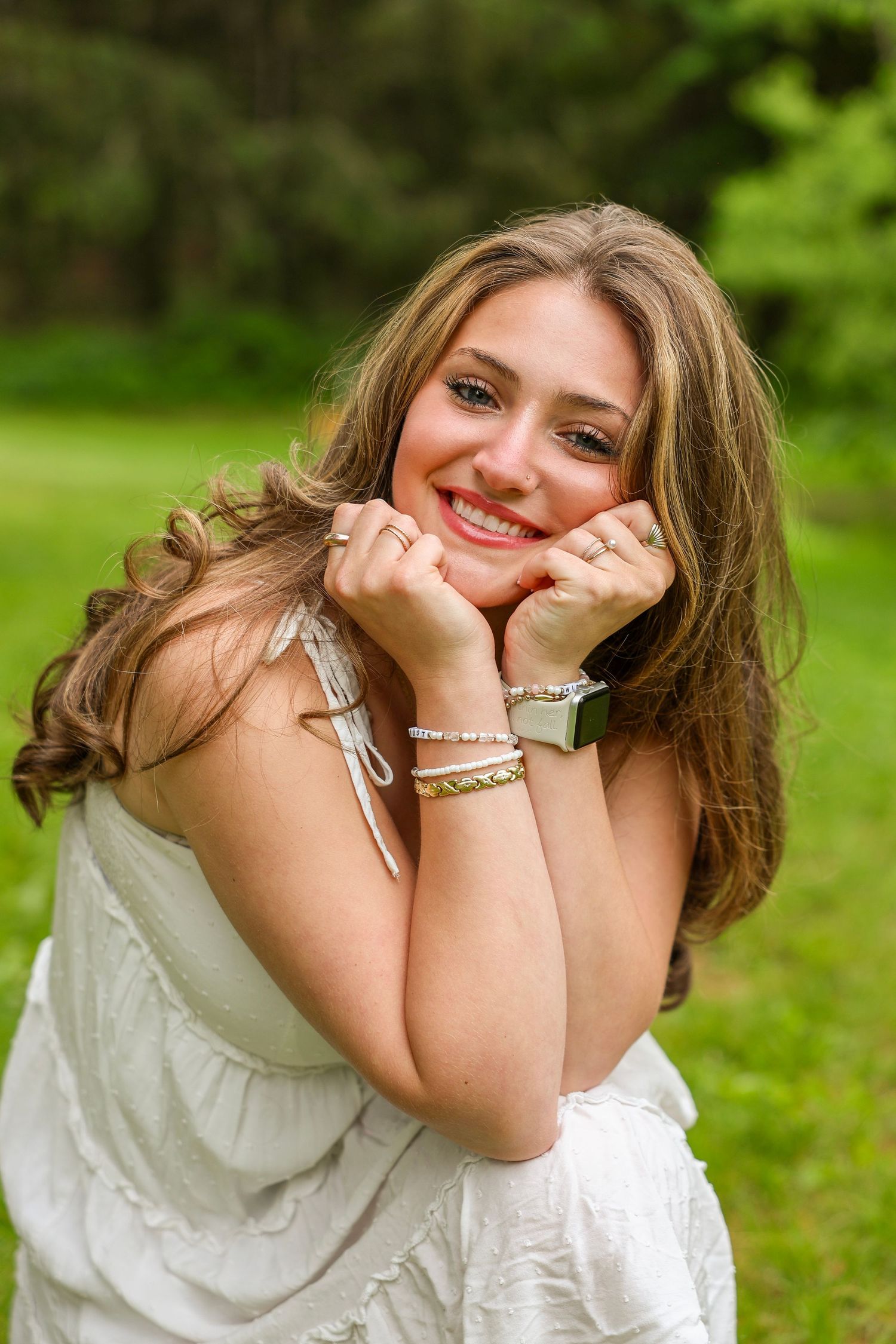 Cheerful young woman with wavy brown hair smiling with hands on chin, sitting outdoors in white dress.