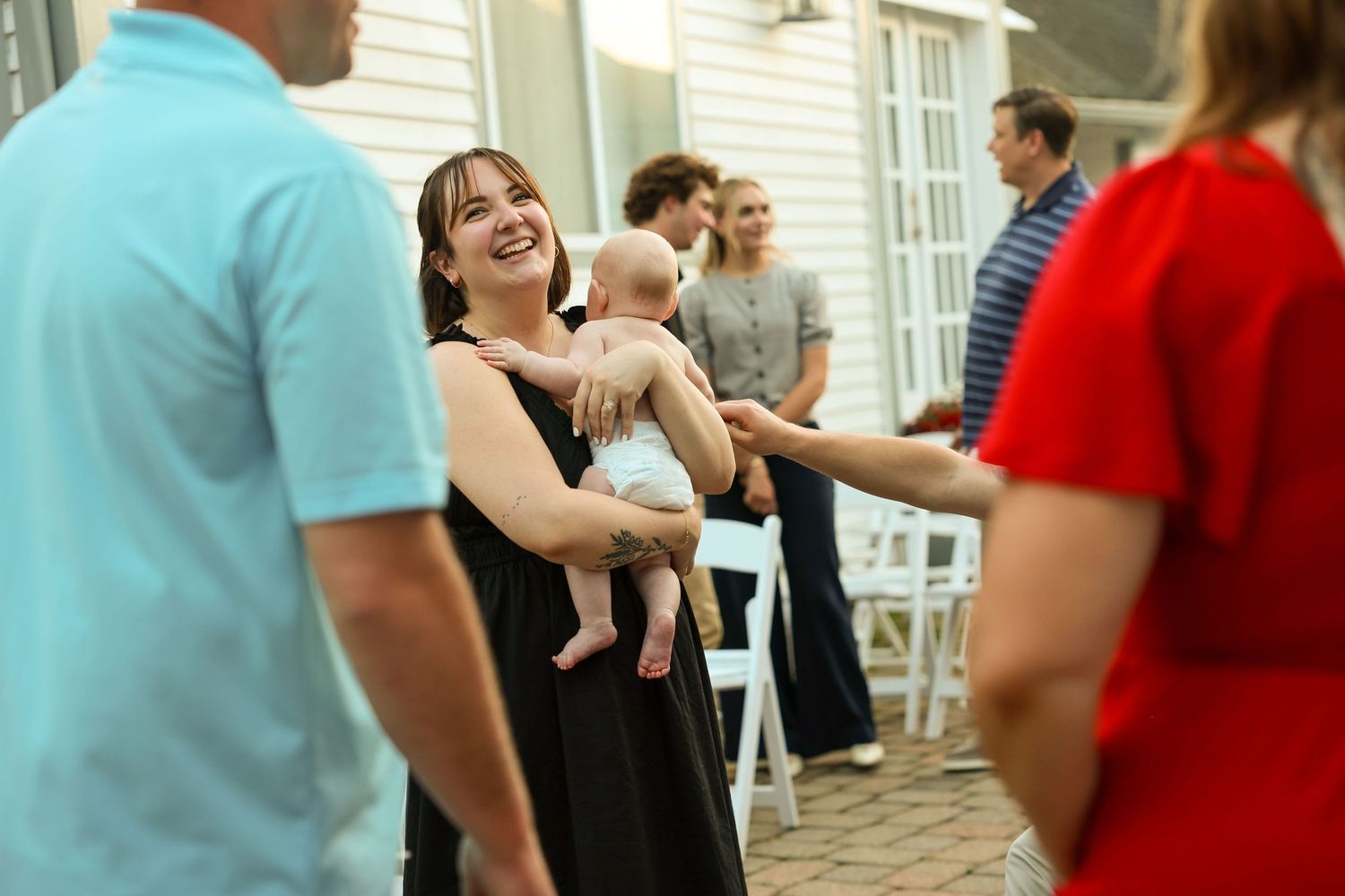Smiling woman in black dress holding baby laughing with guests at an outdoor evening party.