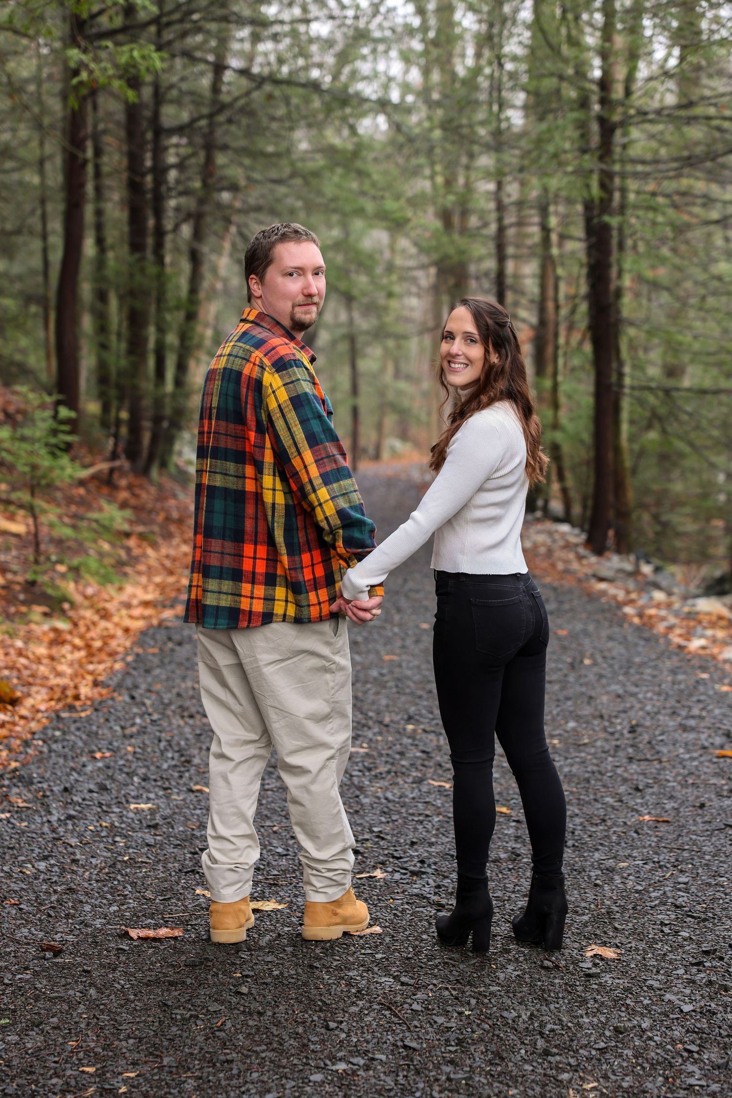 A couple holds hands on a rainy forest path, both looking back at the camera with warm smiles.