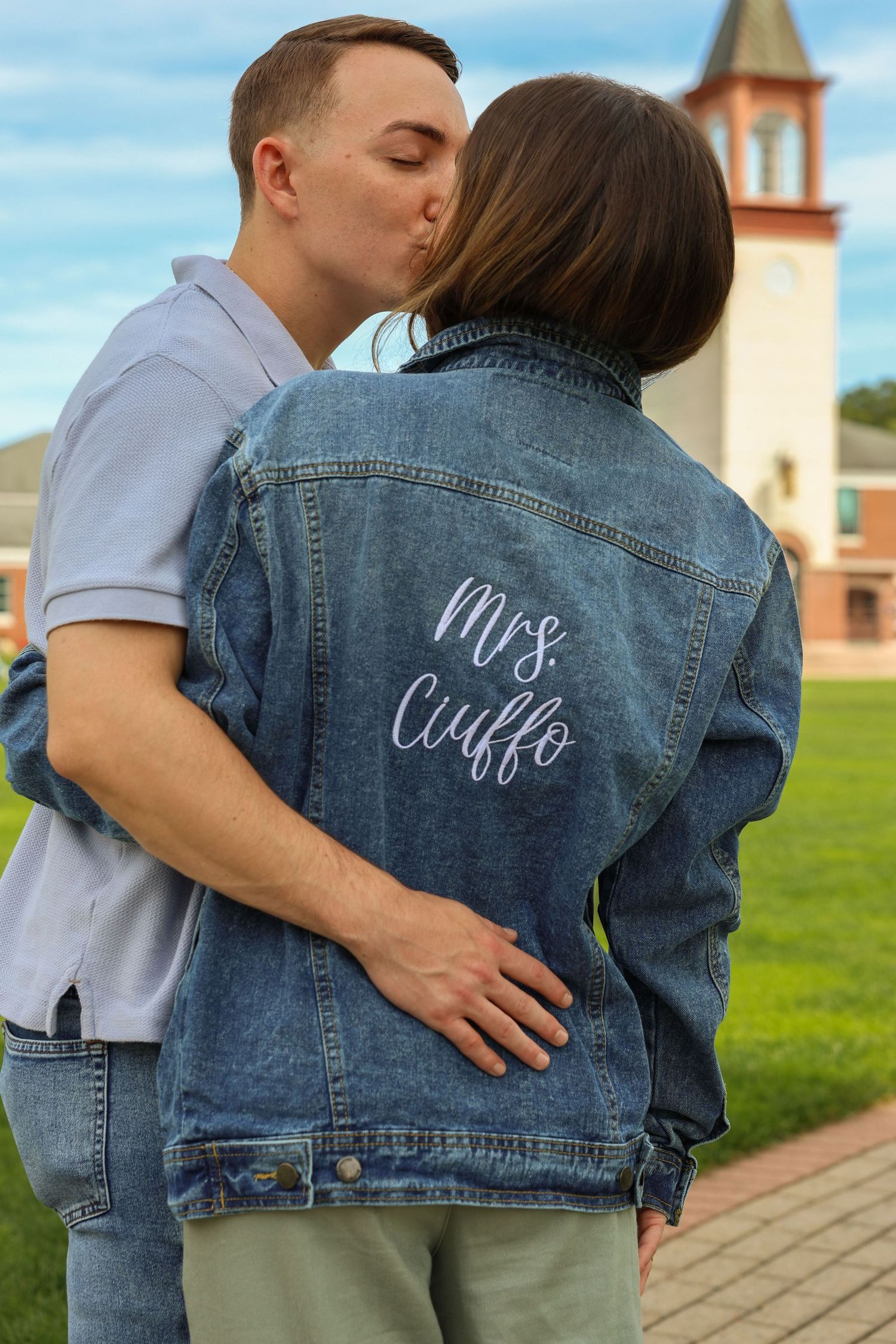 Newly engaged woman in custom Mrs. Ciuffo denim jacket being kissed by her fiance on a college campus.