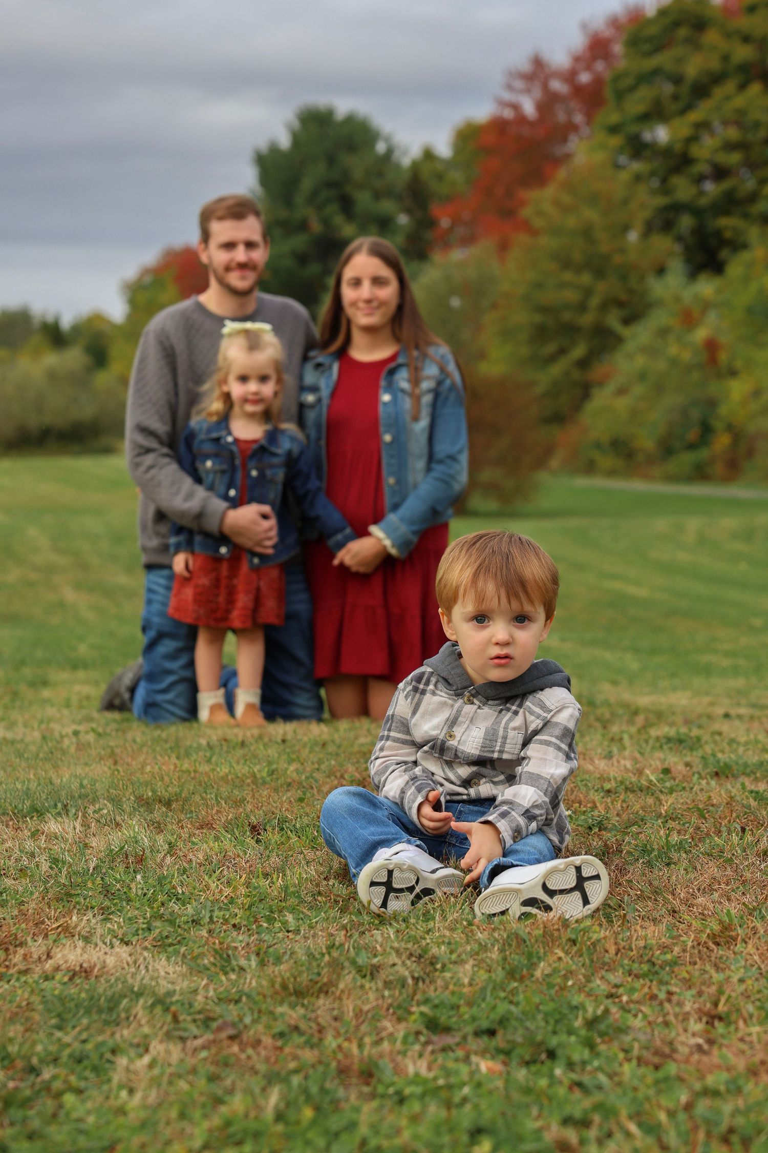 Young toddler sits in foreground as family of four poses in autumn outdoor photo session.