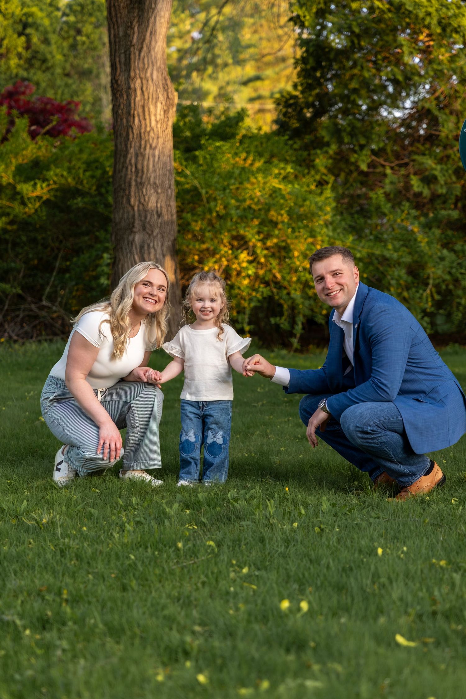 Smiling family of three posing outdoors in park, parents crouching beside standing toddler on green grass.