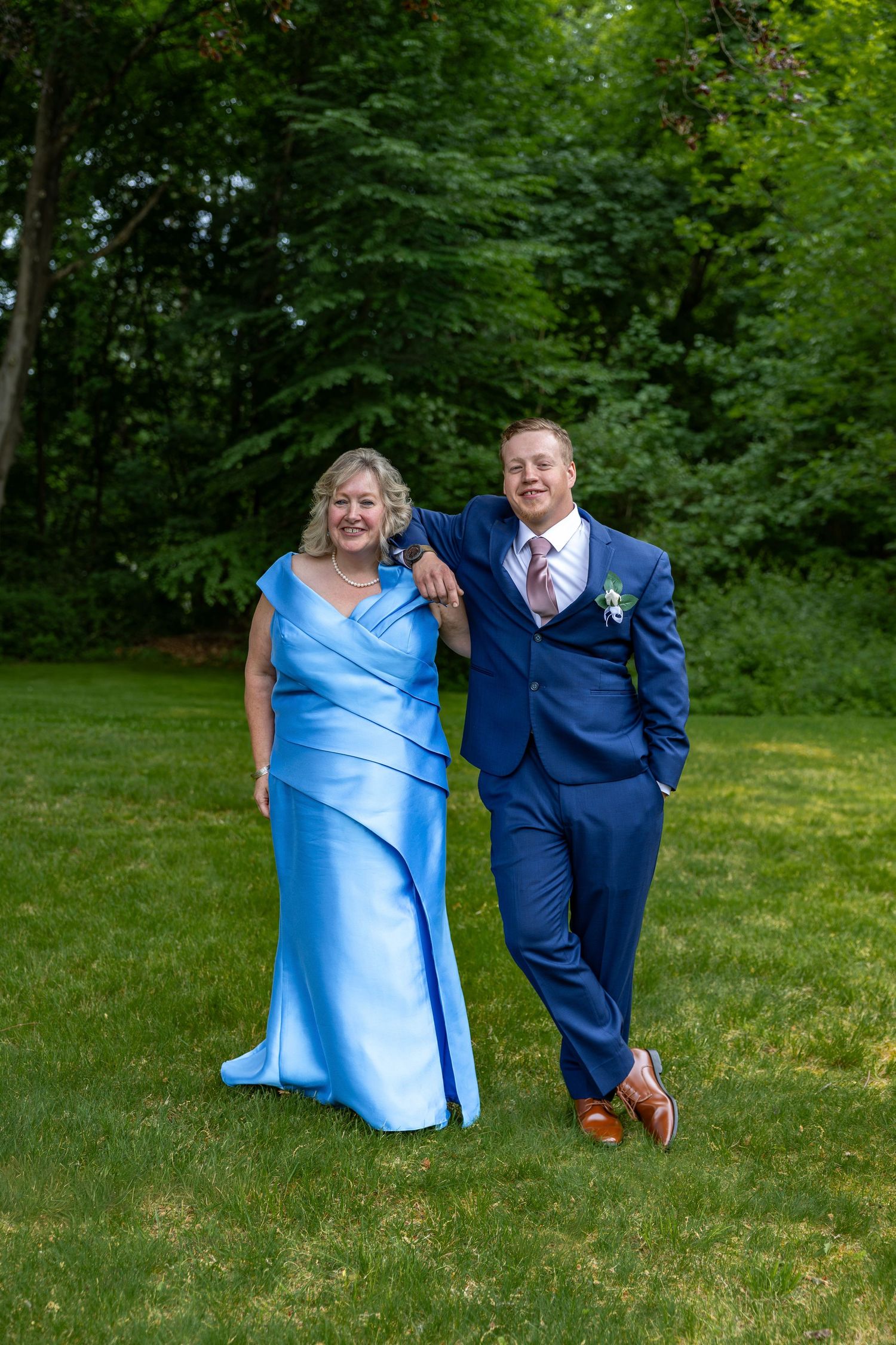 Smiling groom in navy suit with boutonniere poses with woman in light blue formal gown on green lawn.