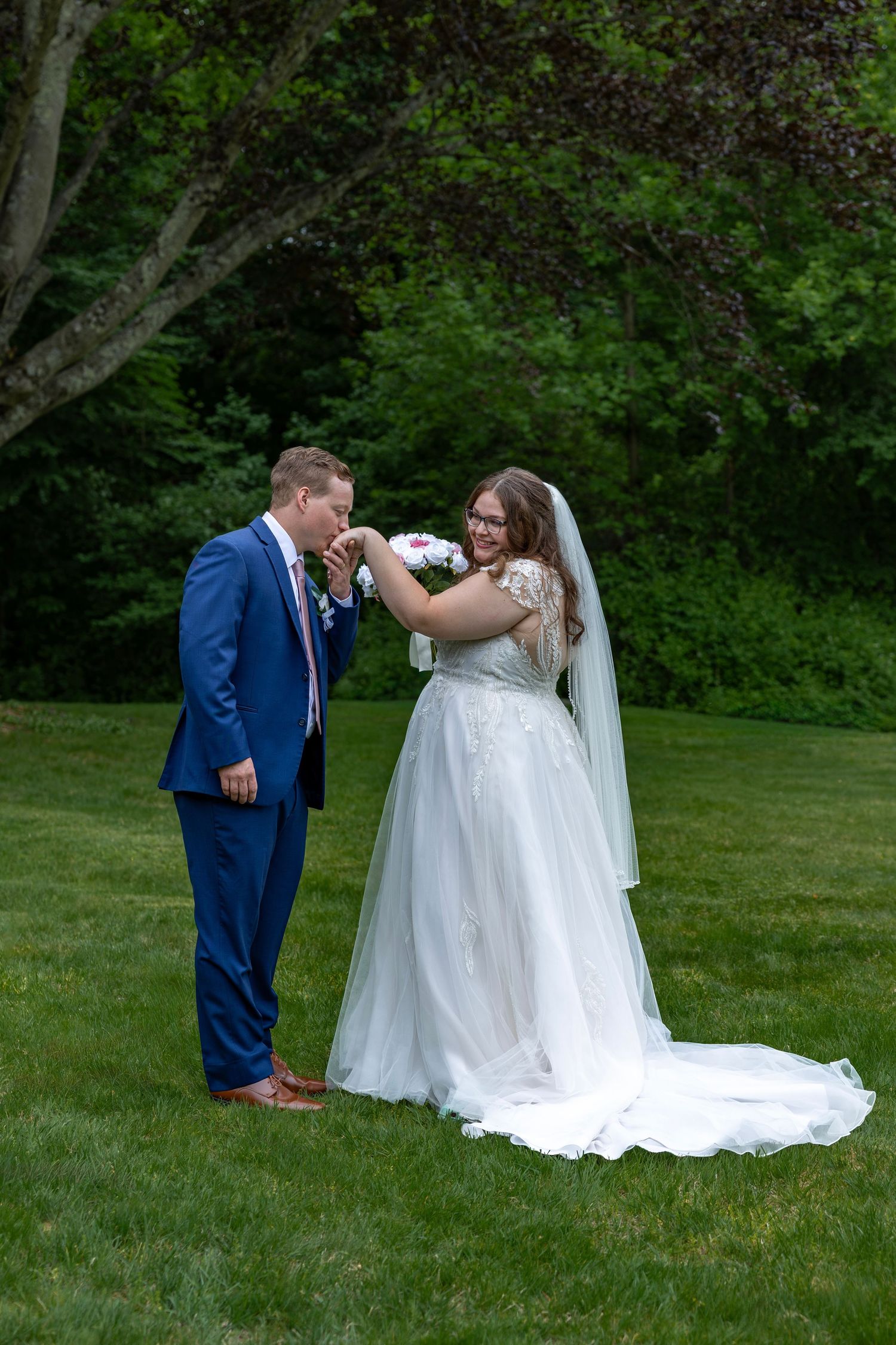 Groom kisses bride's hand as she smiles holding bouquet, standing on green lawn in wedding attire.