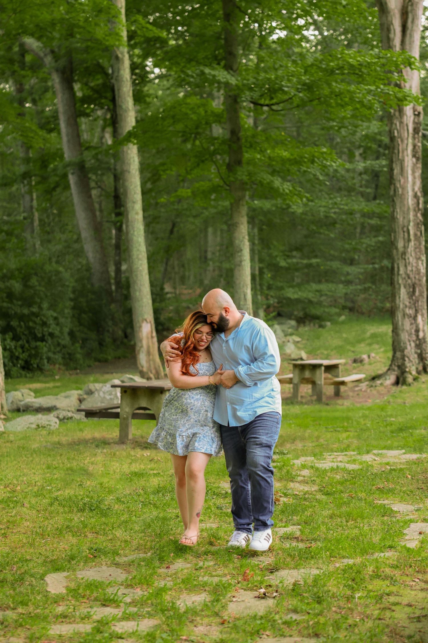 Romantic couple embracing and walking together through a lush green park with tall trees in the background.