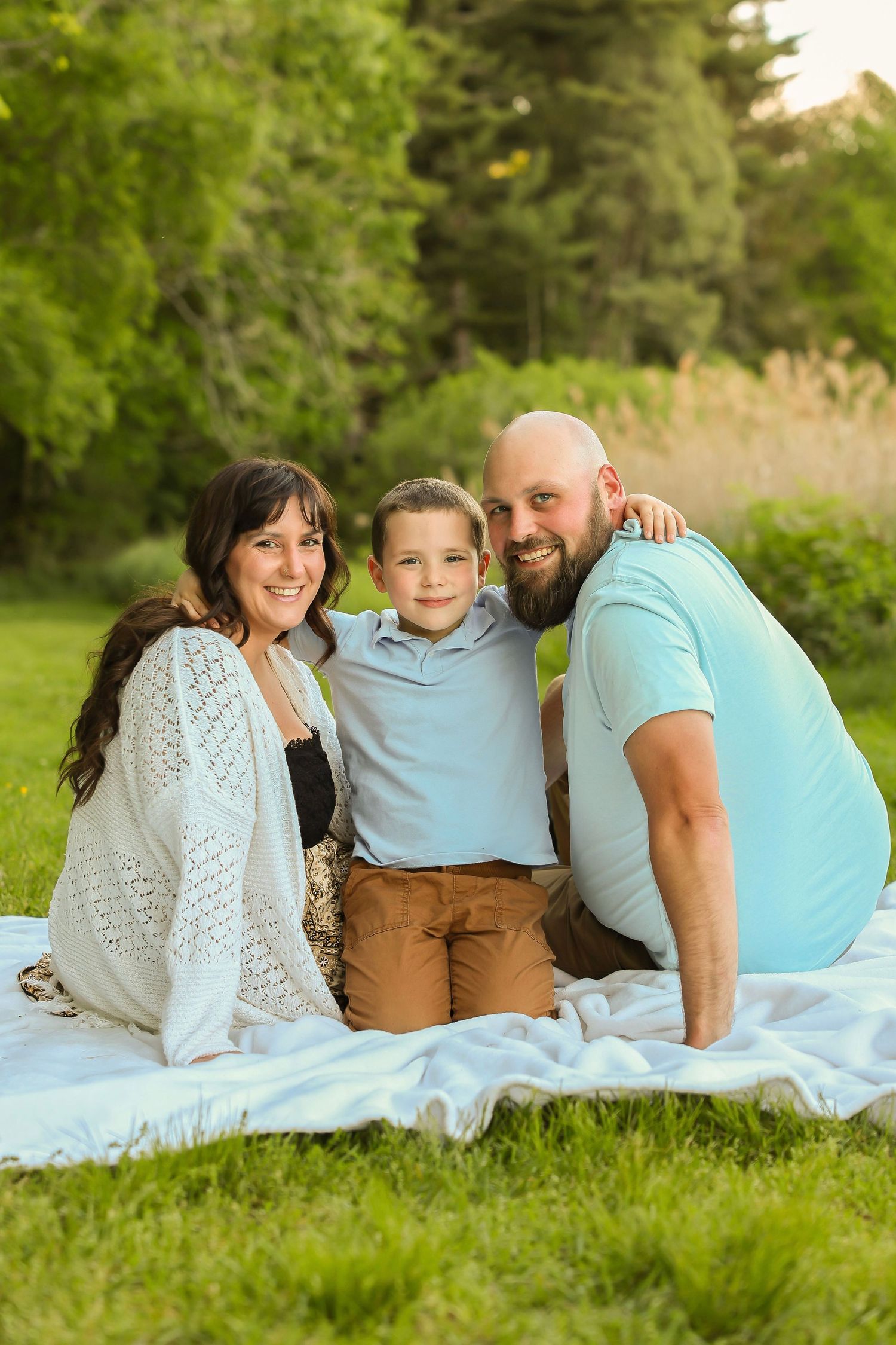 A happy family of three smiles together while sitting on a white blanket on a sunny green lawn.