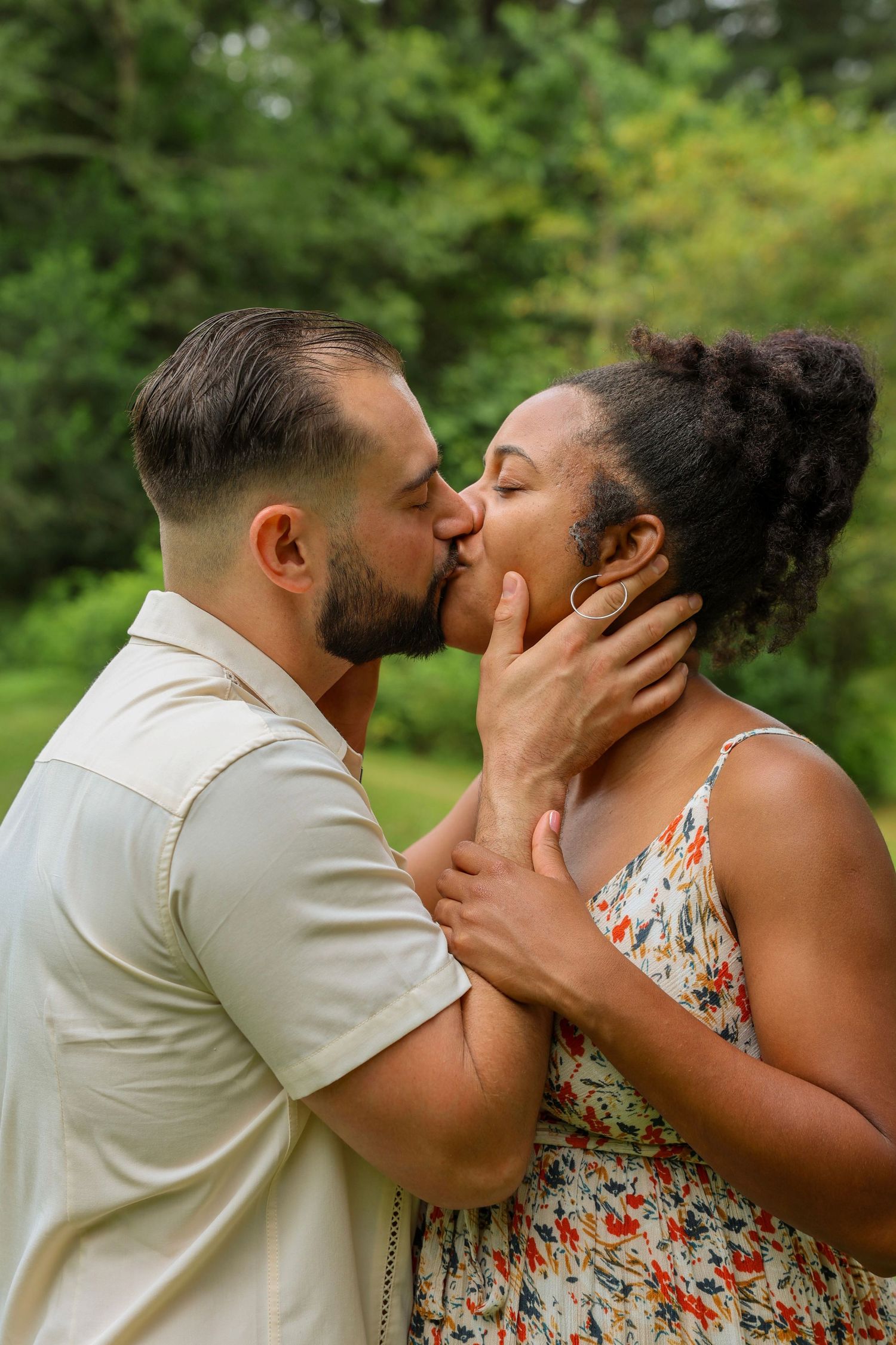 Interracial couple sharing a romantic kiss outdoors surrounded by green trees.