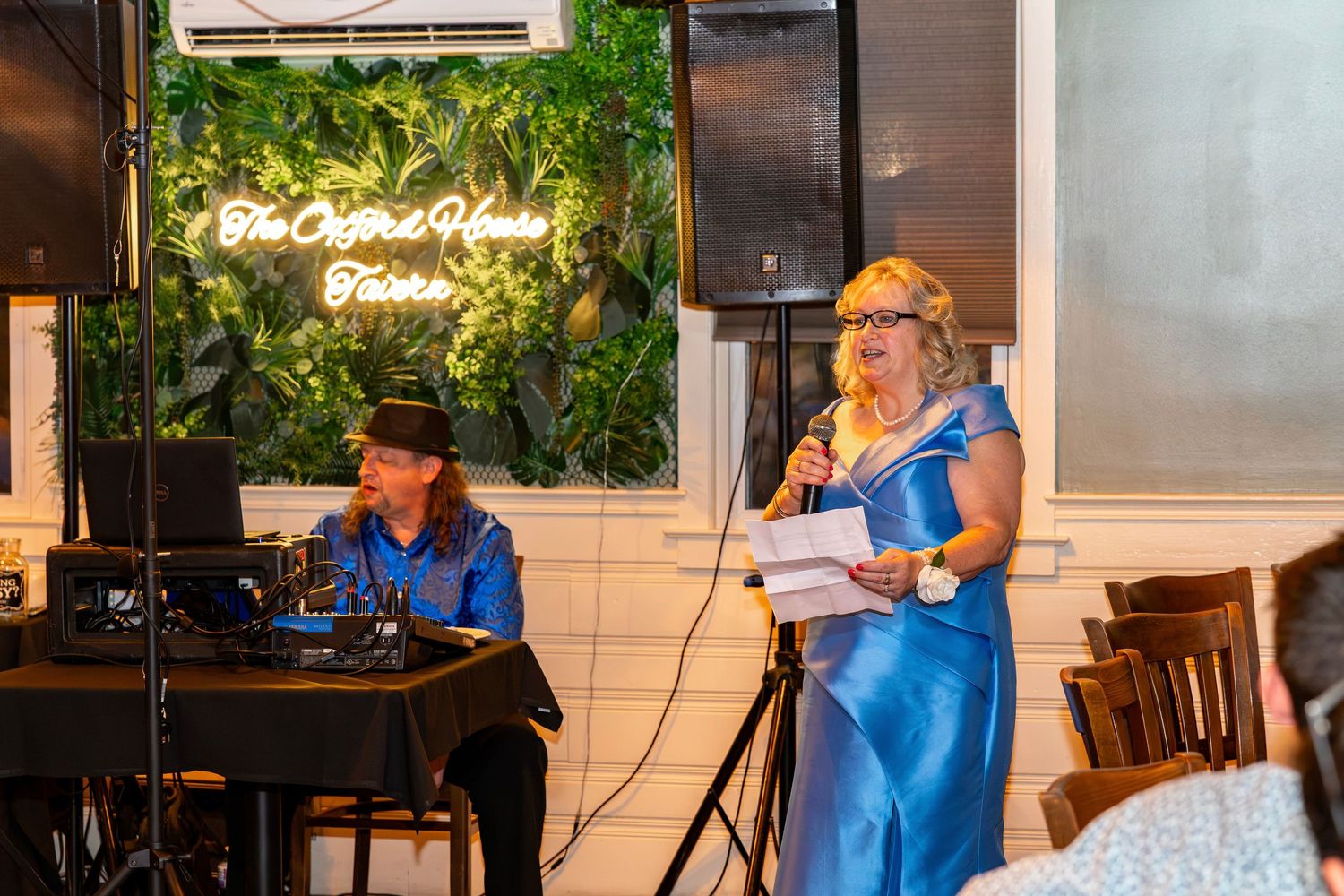 Woman in blue gown gives a speech at wedding reception with DJ performing behind her.