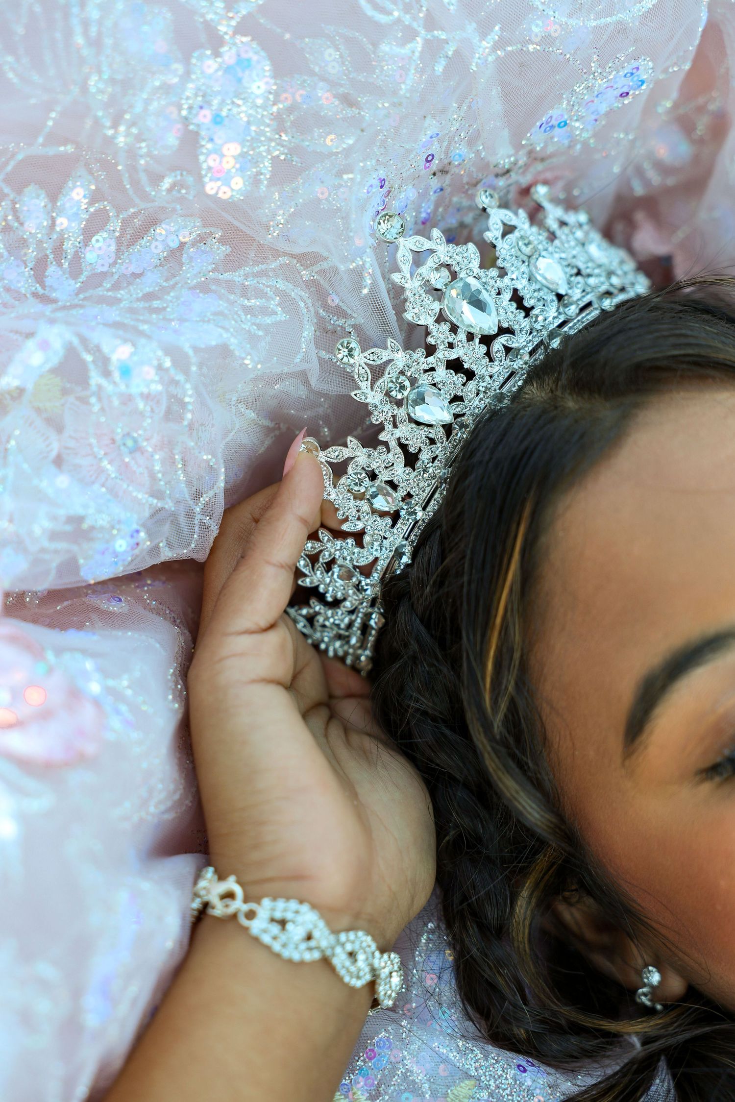 Young woman lying in a pink sparkly quinceañera gown wearing a crystal tiara and diamond bracelet.