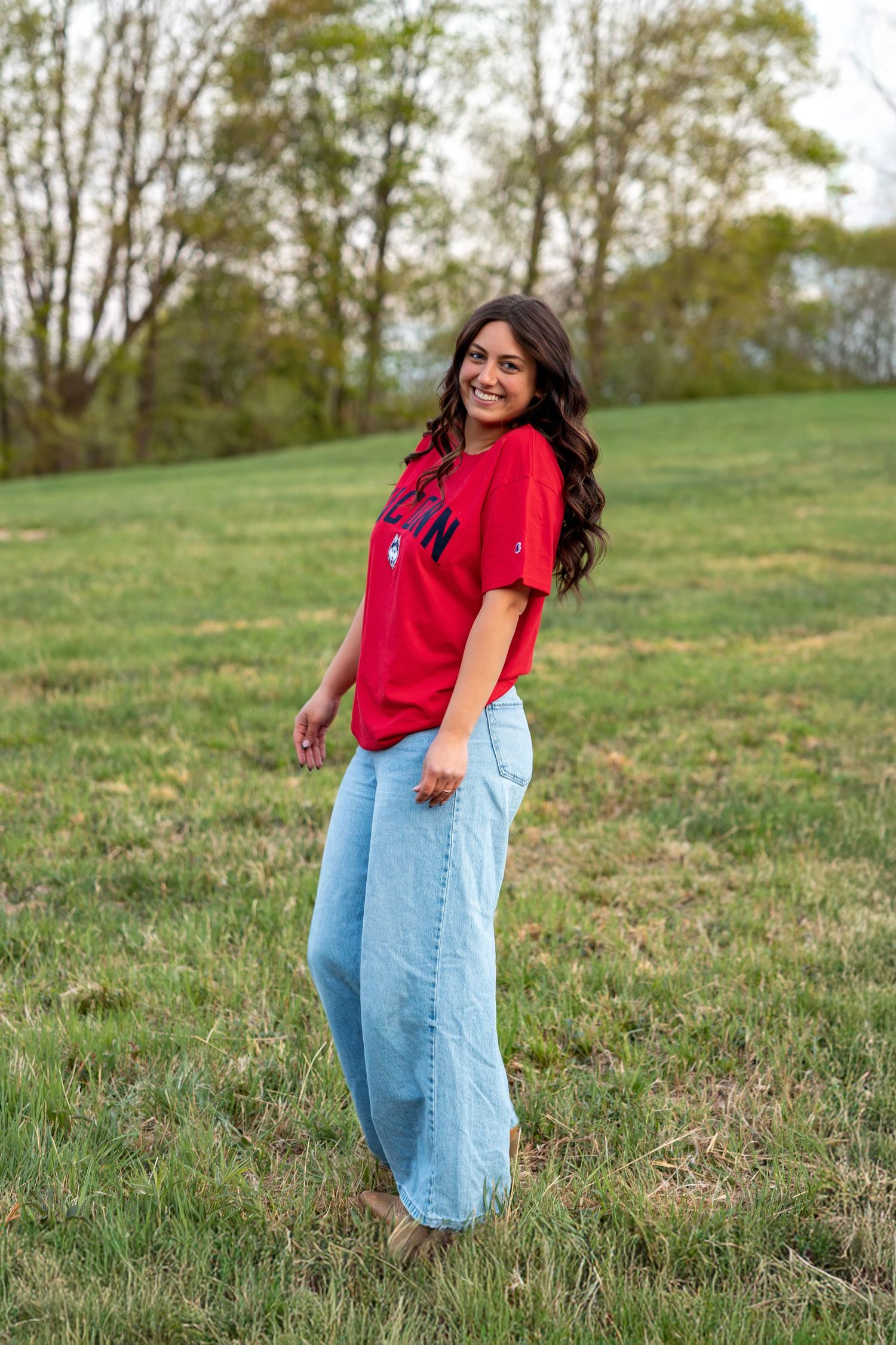 Smiling young woman in red UConn t-shirt and wide-leg jeans posing on green grassy hillside.