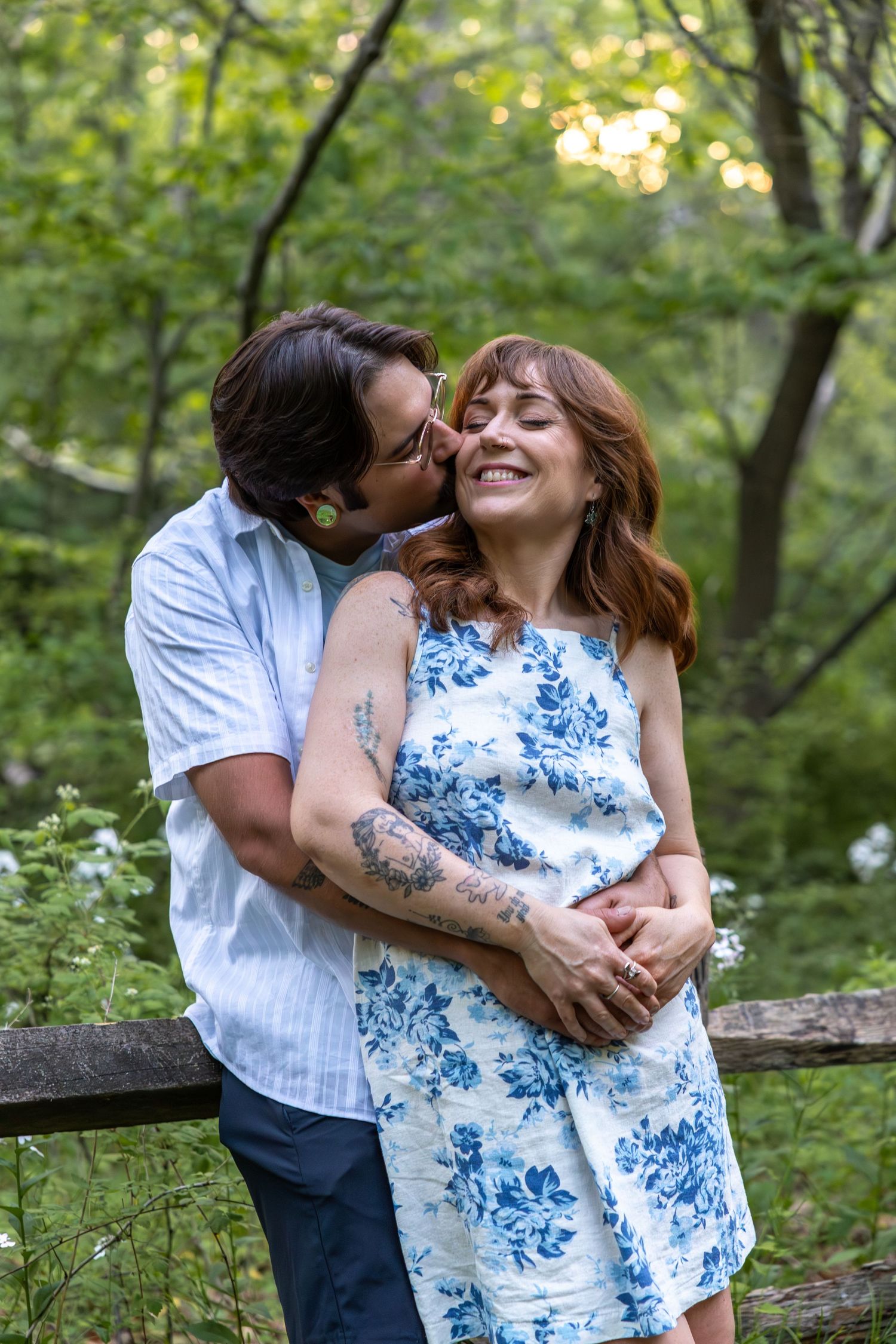 Man kisses woman on cheek while embracing her from behind near a rustic fence in a green forest setting.