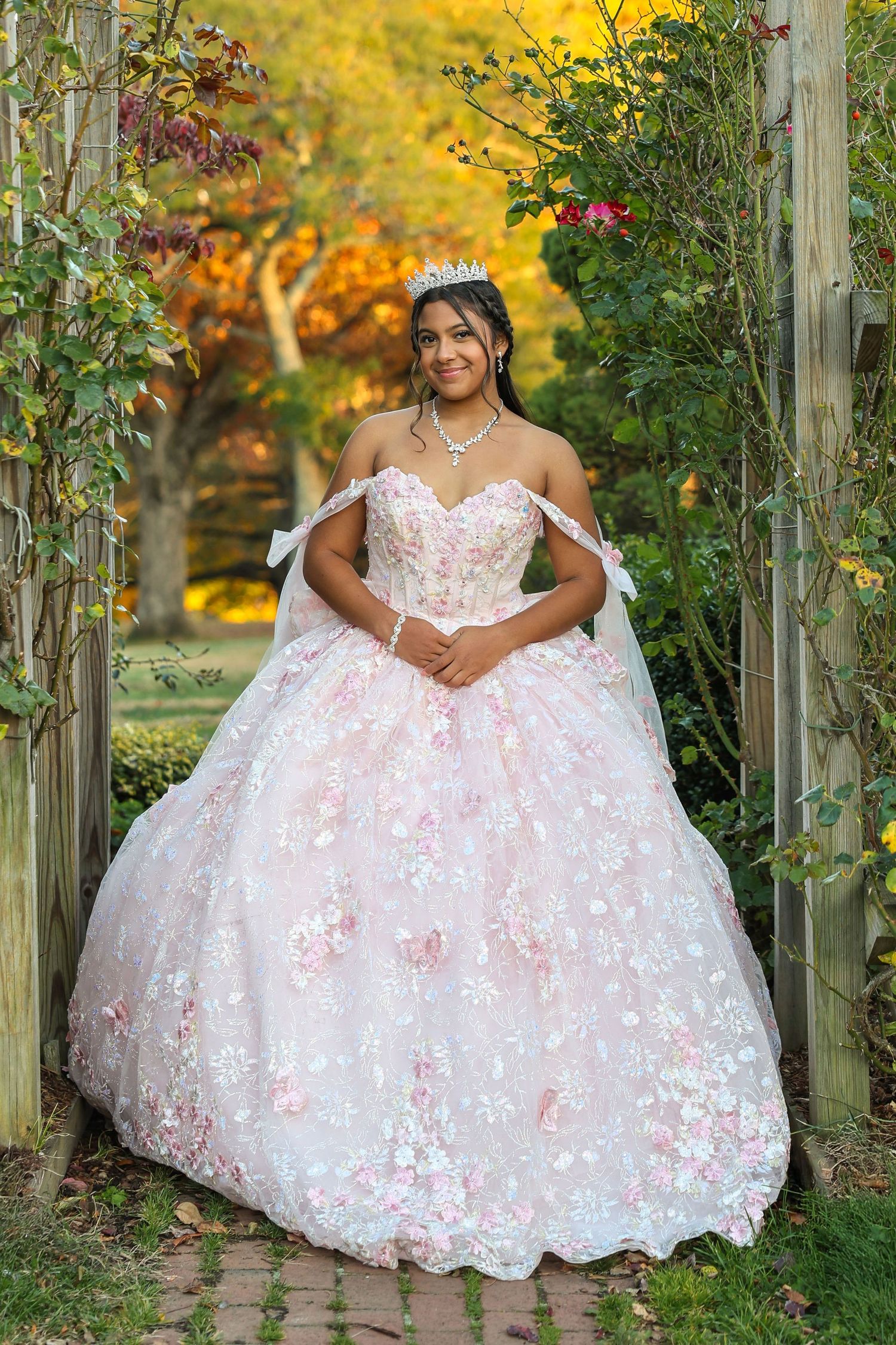 Young woman in pink floral quinceañera ball gown standing under wooden garden arbor in autumn.