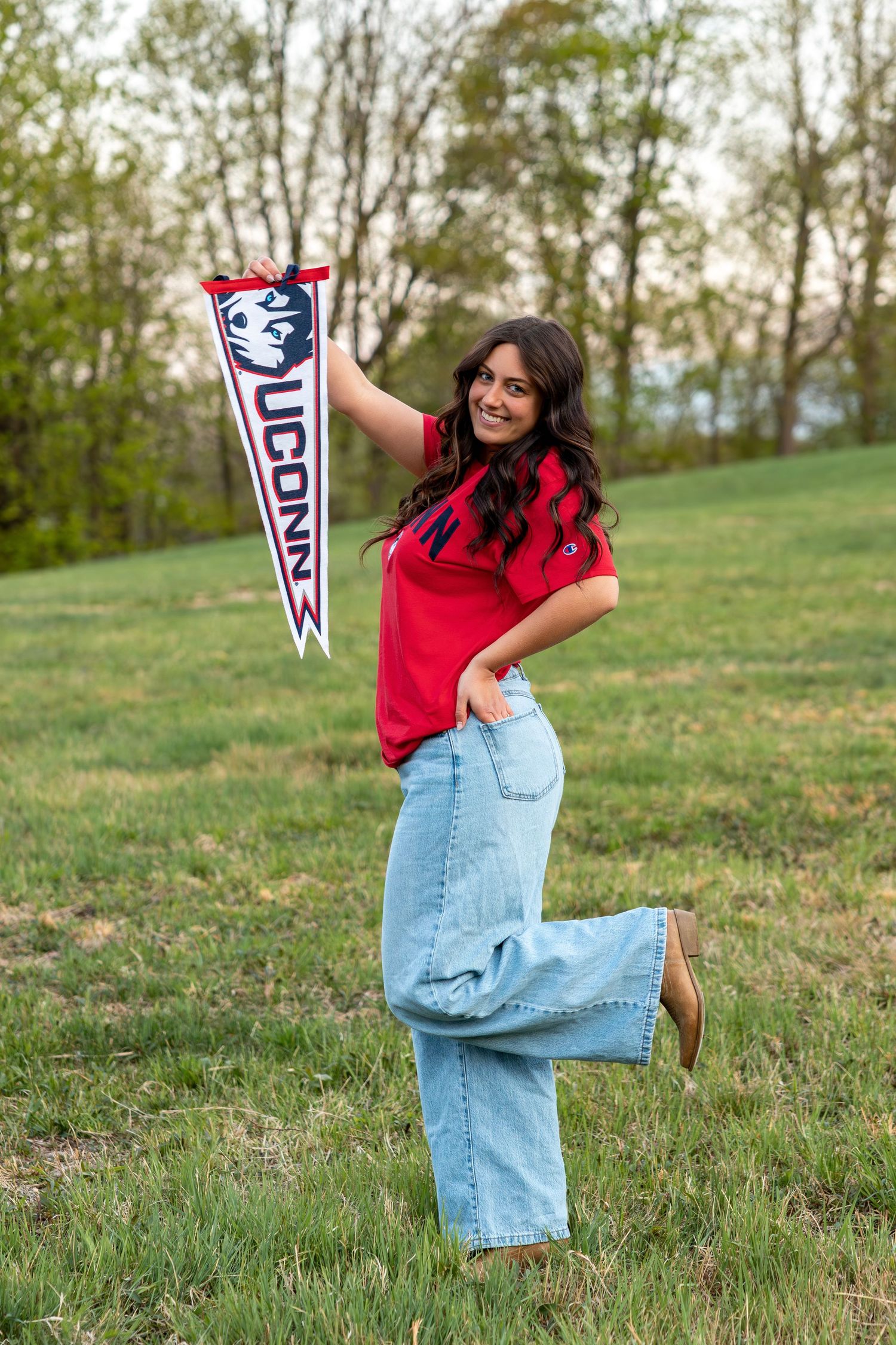 Playful young woman raising UConn pennant flag in grassy field wearing red tee and wide-leg jeans.