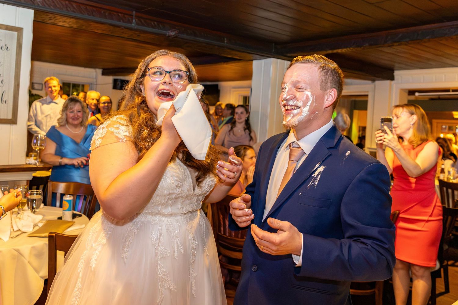 Bride wipes cake from her face as groom laughs with frosting on his cheek at reception.