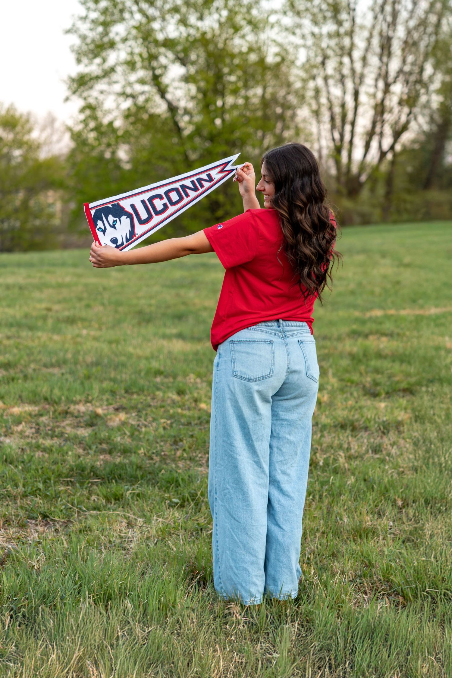 Young woman holding UConn Huskies pennant flag in green field wearing red tee and wide-leg jeans.