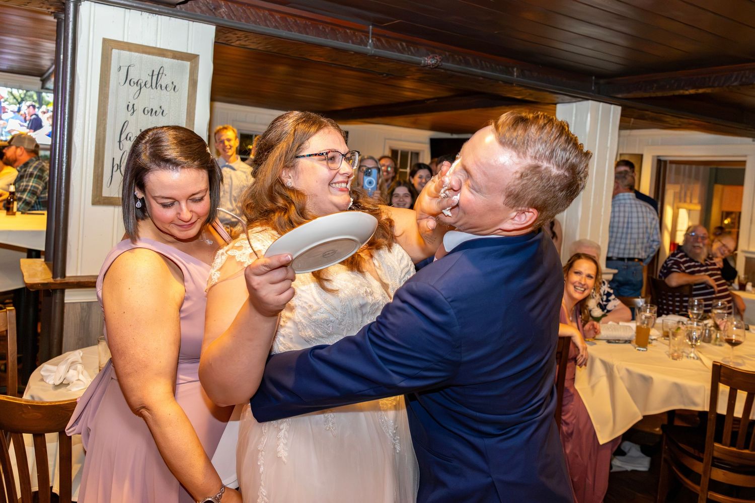 Bride smashes wedding cake into groom's face as bridesmaid laughs at indoor reception.
