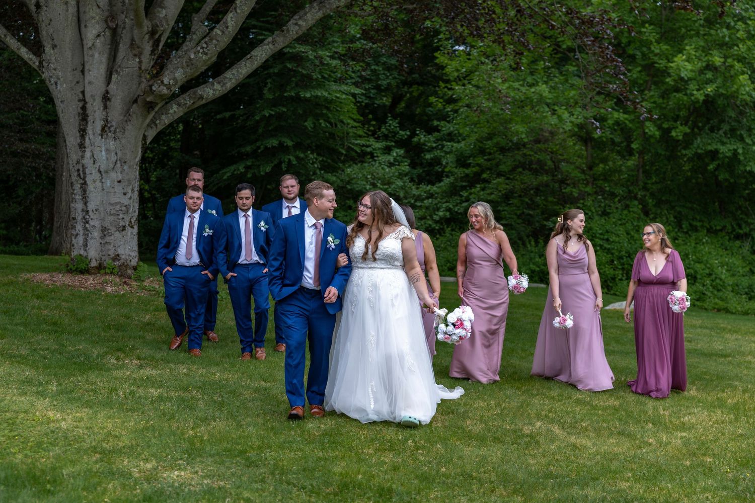 Wedding party walks across green lawn with bride in white gown and bridesmaids in mauve dresses.
