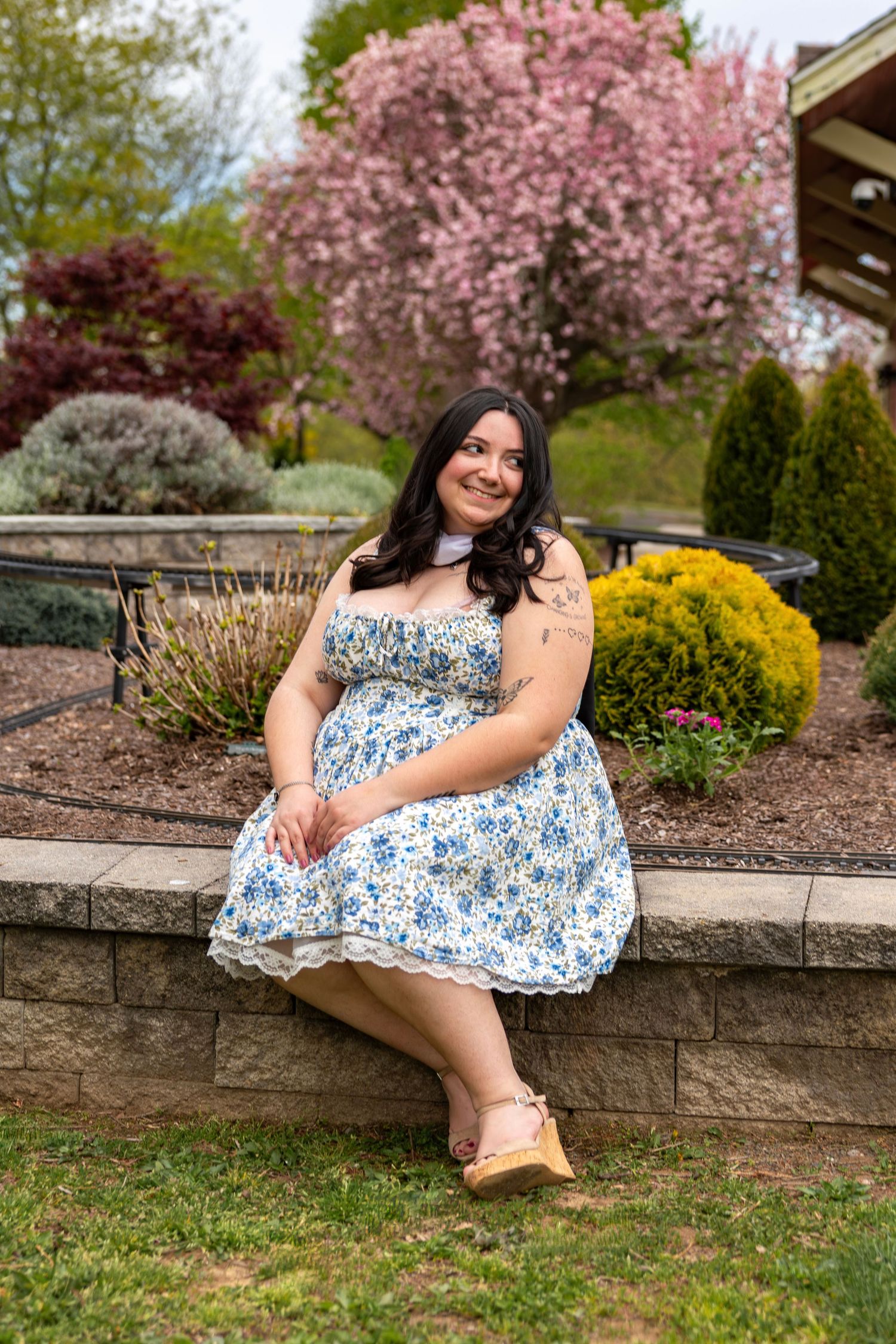 Smiling young woman in blue floral dress sits on stone garden wall surrounded by colorful spring blooms.